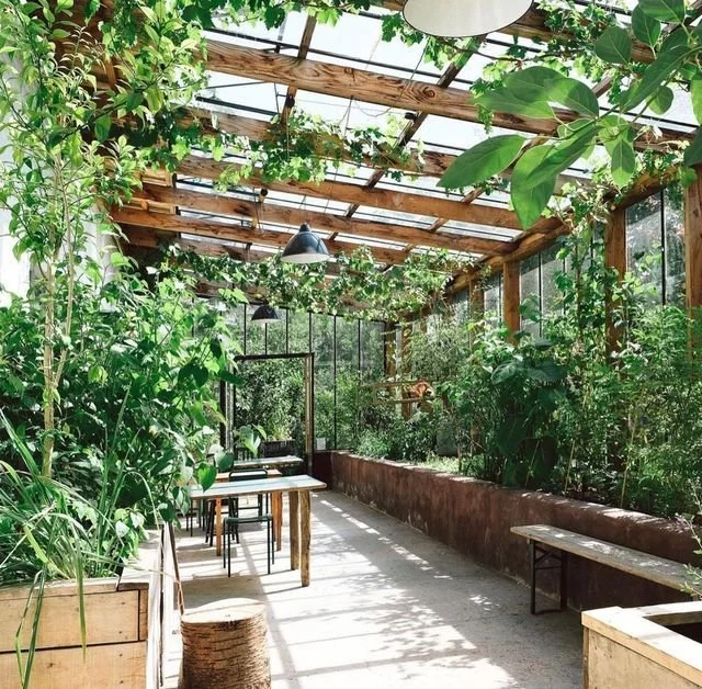 Binnen een kas of serre met veel groene planten en natuurlijke houten structuren, tafels en stoelen, voorzien van glas en metalen lampen.