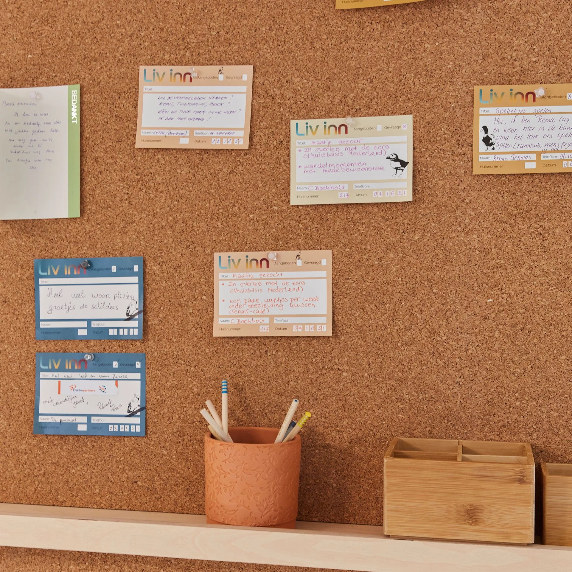 Corkboard met diverse kaartjes met tekst en tekeningen, een oranje pot met pennen en twee houten opbergdozen.