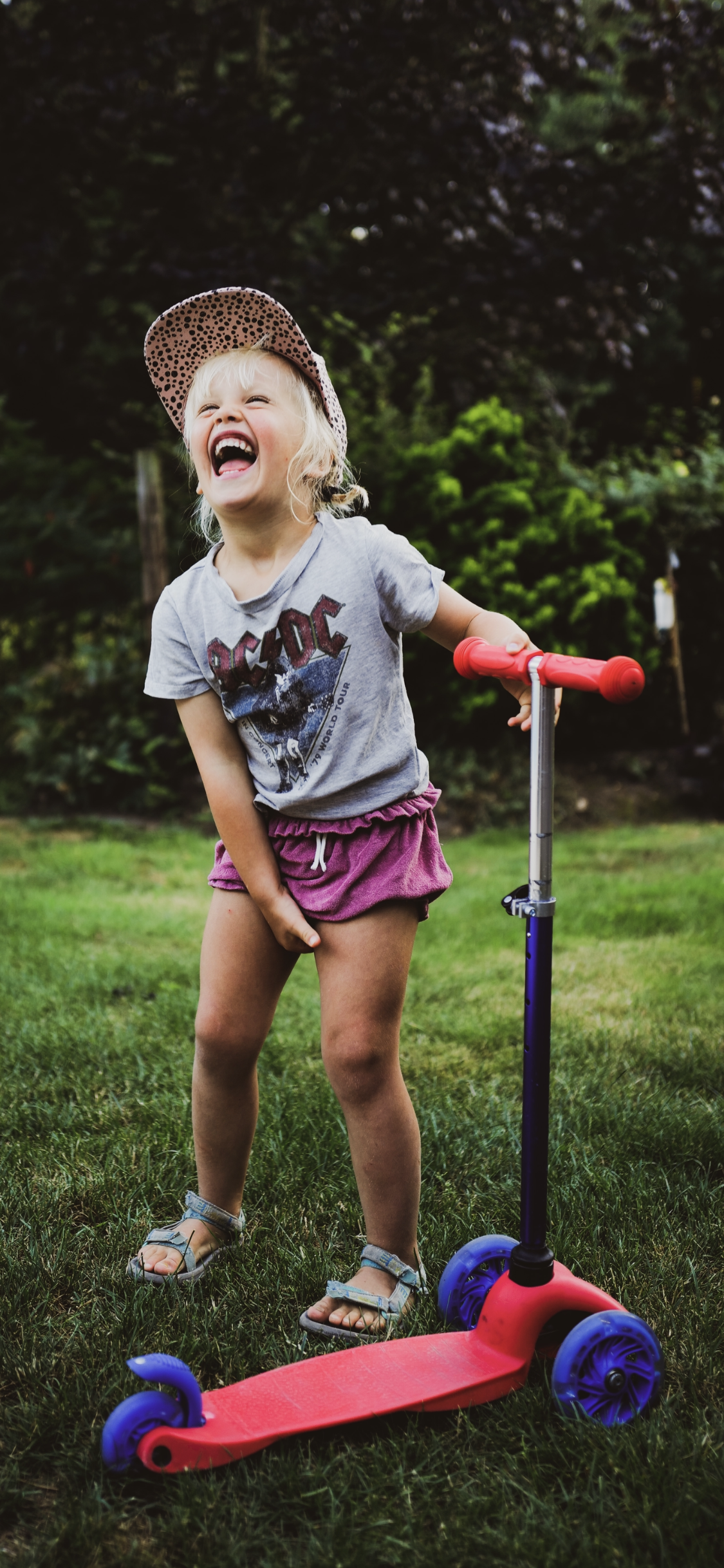 Een lachende jonge meisje met een roze pet, een grijze AC/DC T-shirt en roze shorts, staat buitenshuis op gras met een skate of skelter, terwijl ze plezier heeft.