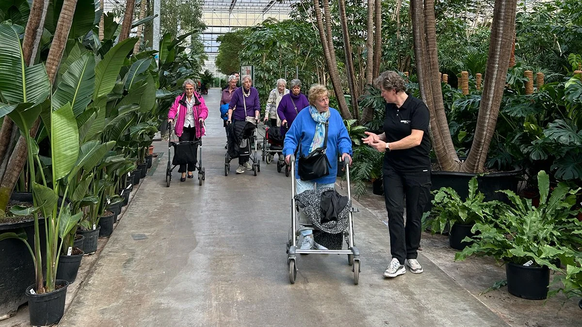 Vijf oudere vrouwen met wandelstokken en rollators wandelen door een kas met grote groene planten, terwijl een jonge vrouw hen aanspreekt.