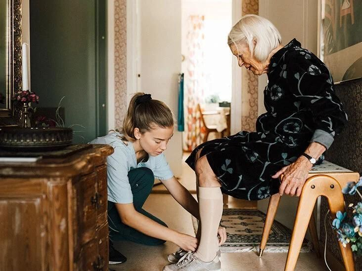 Een jonge vrouw helpt een oudere vrouw met haar schoen in een huiselijke omgeving.
