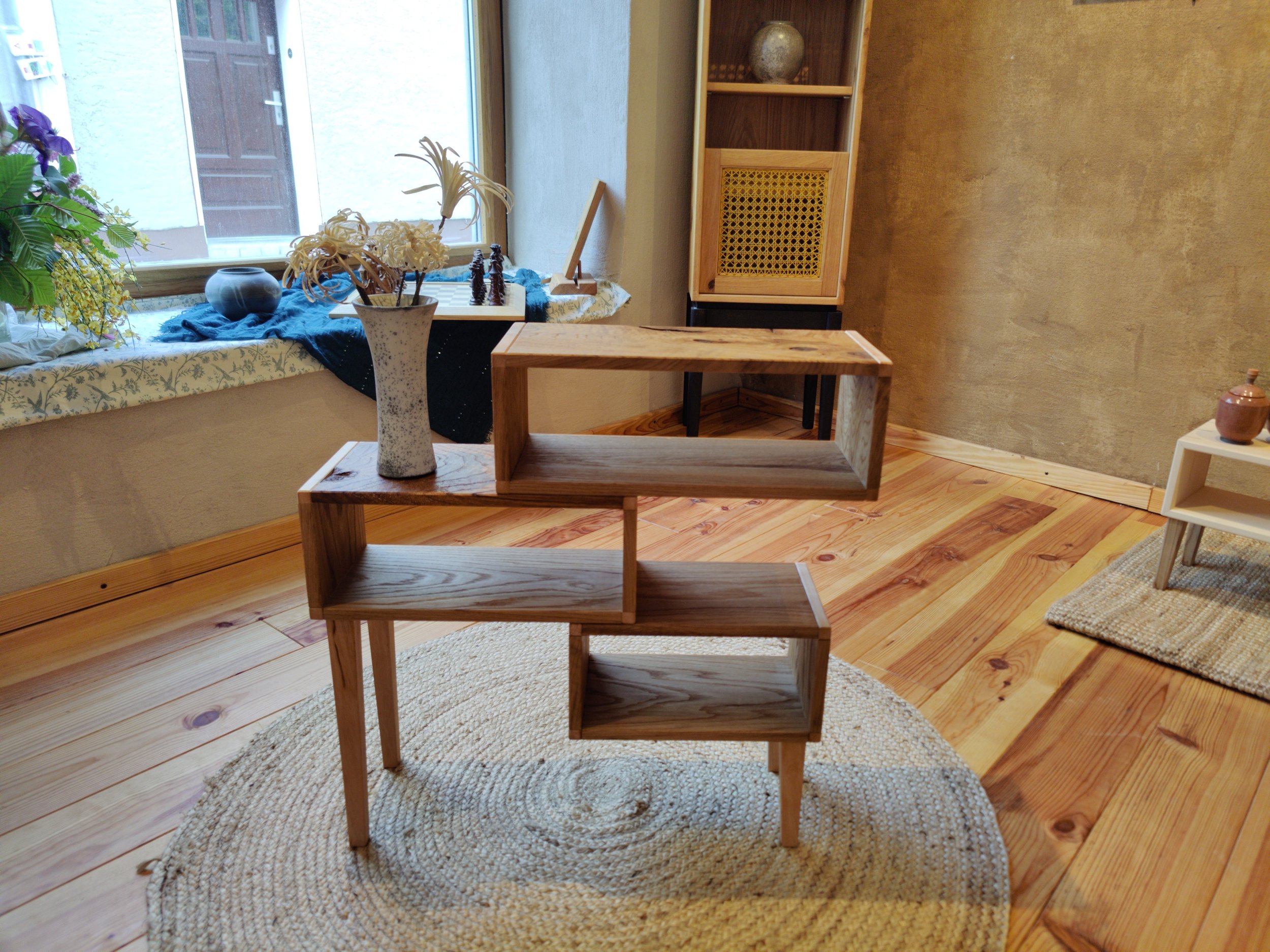 Ein Raum mit Holzboden, einem runden Teppich, einem Holzregal und einer Fensterbank. Es gibt eine Vase mit getrockneten Blumen auf einem anderen Regal und einige Dekorationsgegenstände. Das Licht kommt vom Fenster auf der linken Seite.