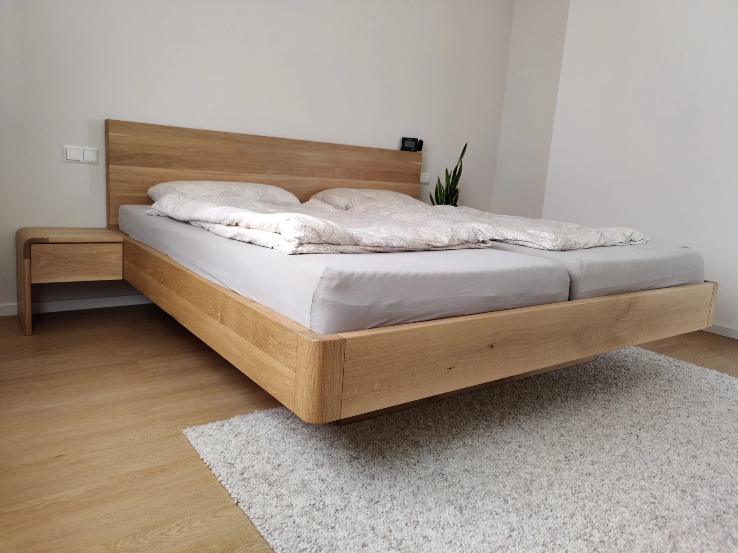 Ein modernes Schlafzimmer mit einem großen Holzbettenrahmen, weißen Bettlaken, zwei Kissen und Pölstern, ein kleines Nachttisch auf der linken Seite, eine Pflanze im Hintergrund und eine beige Gardine im Hintergrund.