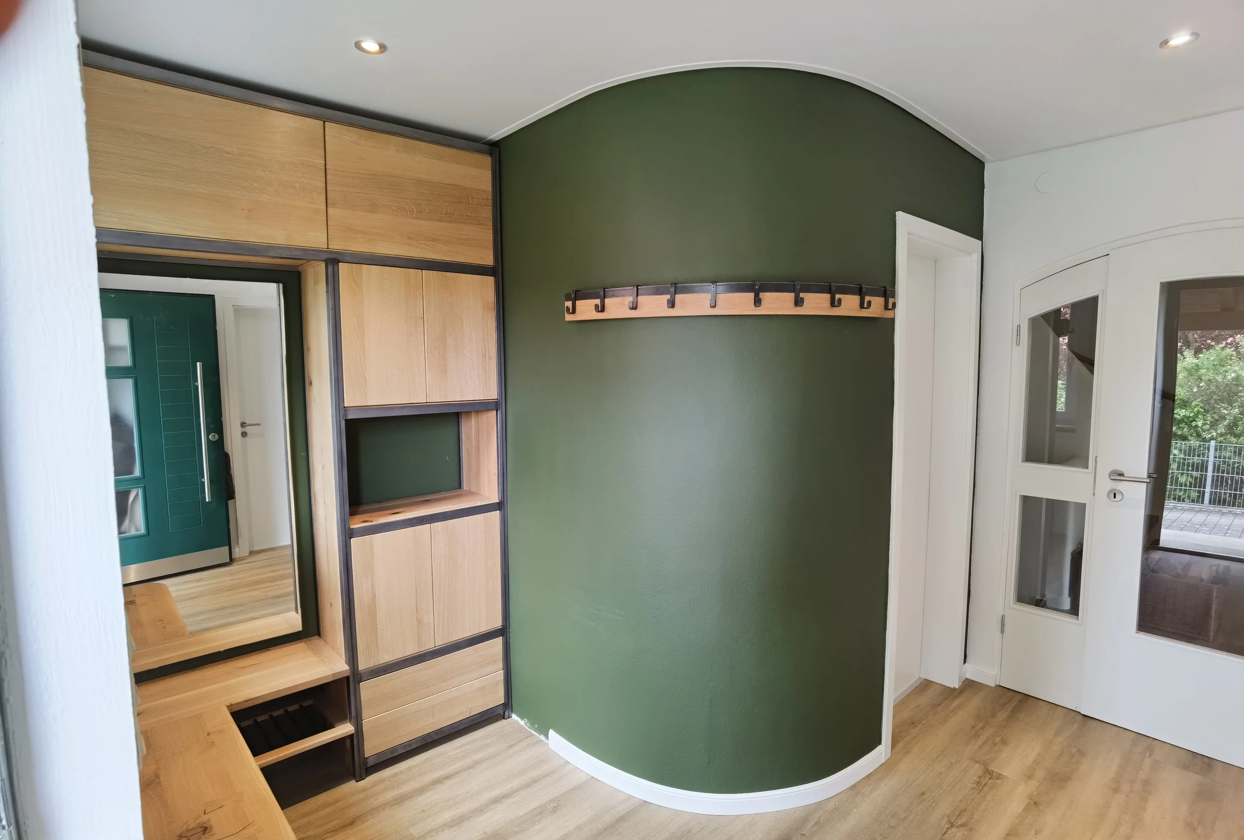 Innenaufnahme eines Flurs mit einer grünen Wand, einem Holzpaneel mit Haken, weißen Türen und einem großen Spiegel.