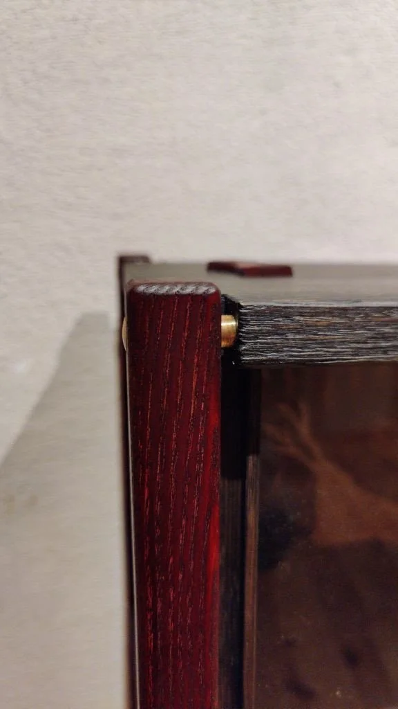 Nahaufnahme eines Holzrahmens mit einem transparenten Glasfenster, rote Ecken und einer goldfarbenen Schraube.