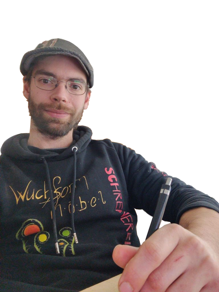 Ein Mann trägt eine dunkle Kapuzenjacke und eine Mütze, sitzt an einem Tisch und schreibt mit einem Stift.