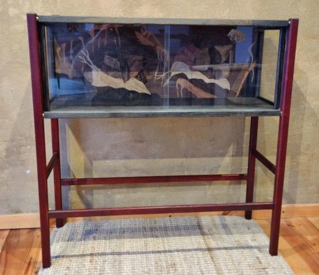 Ein schwarzer, rechteckiger Glaskasten auf einem roten Metallgestell, steht auf einem Holzfußboden vor einer beige Wand, auf einer kleinen Rolle befindet sich eine bunte Decke mit Streifenmuster.