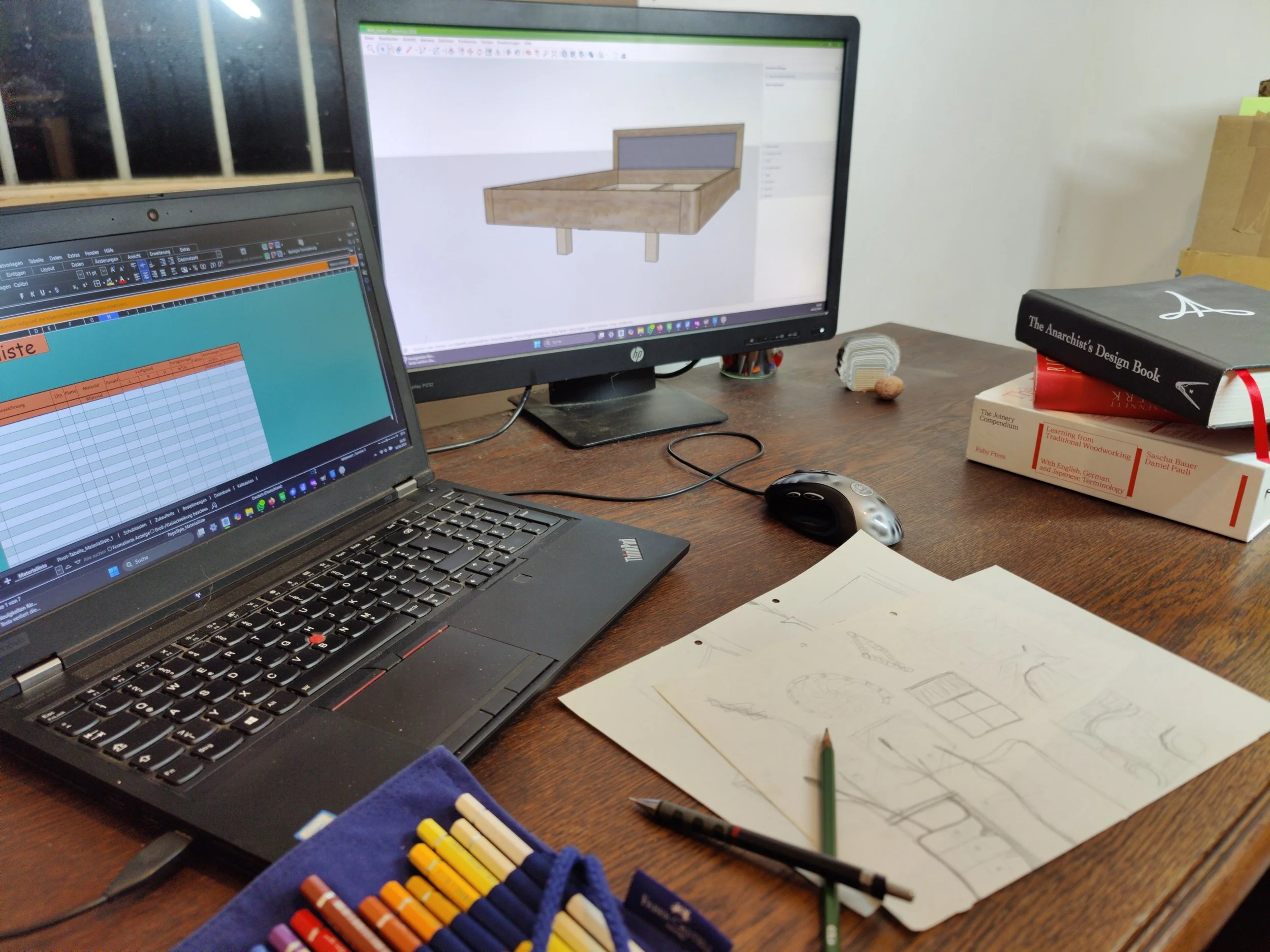 Schreibtisch mit two Monitoren, einem Laptop, Skizzen, Buntstiften, und Büchern.