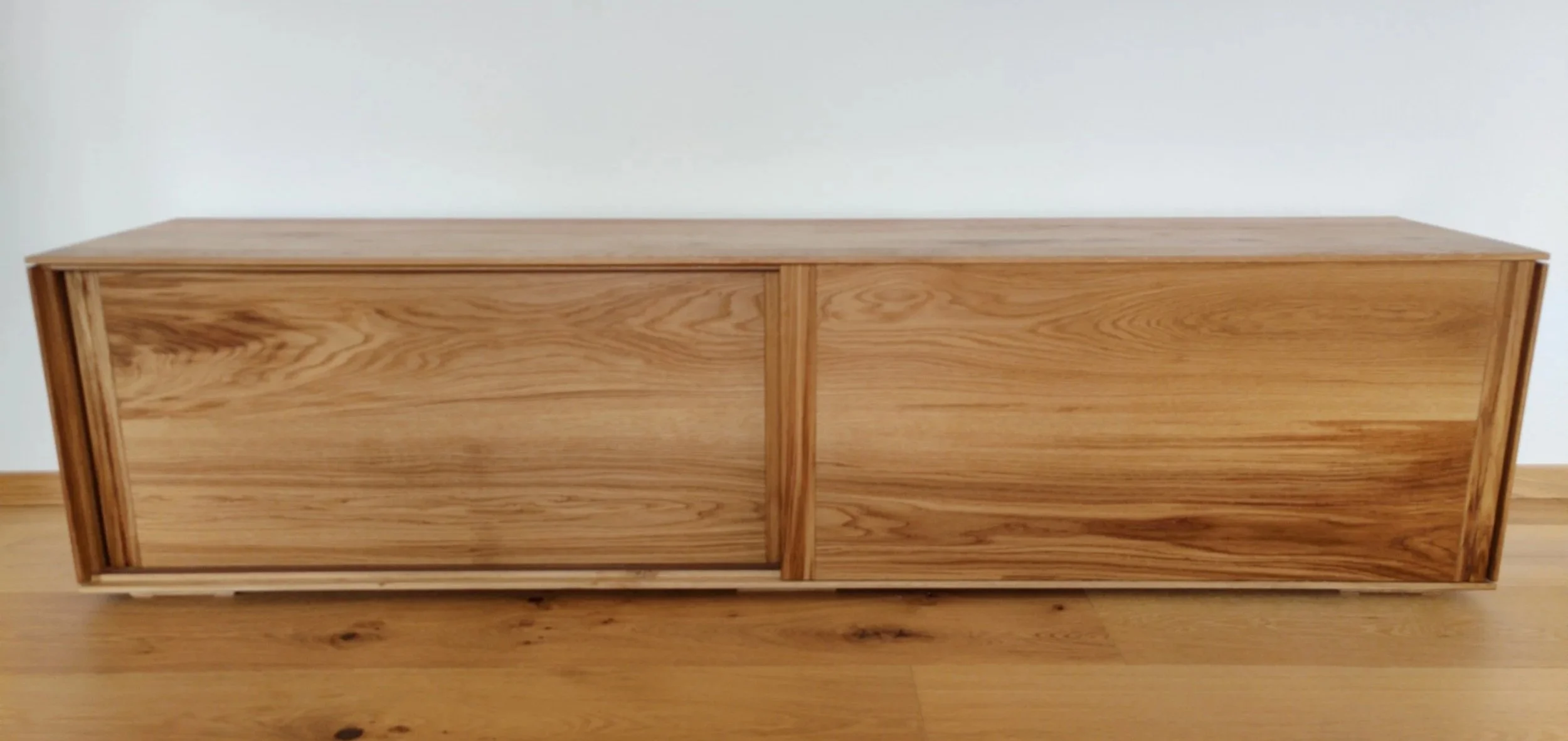 Ein rechteckiger Holztisch mit zwei Schiebetüren, aus hellem Holz gefertigt, steht auf einem Holzboden vor einer weißen Wand.