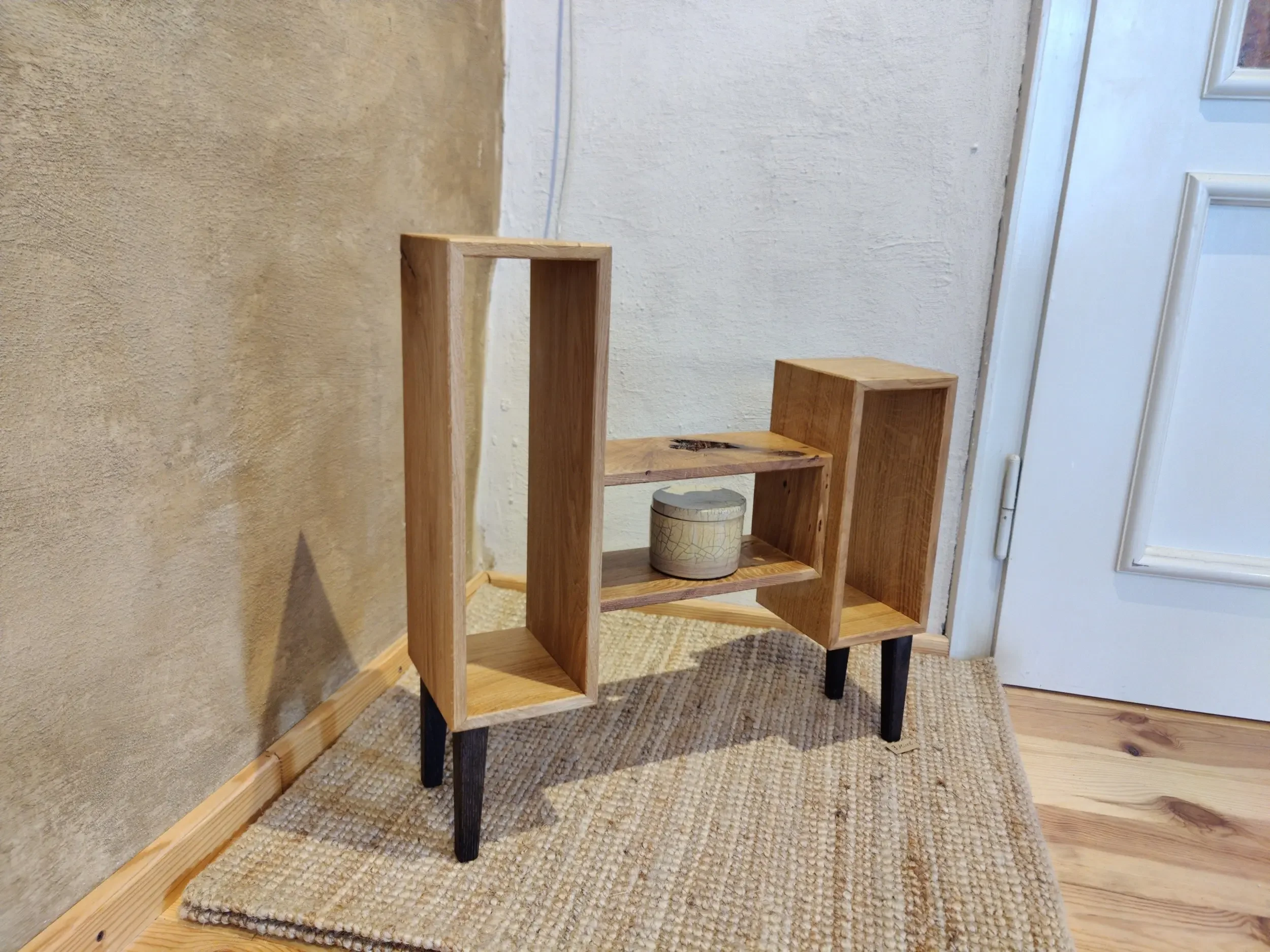 Ein modernes Holzregal mit schwarzen Beinen, auf einem Teppich neben einer weißen Tür.
