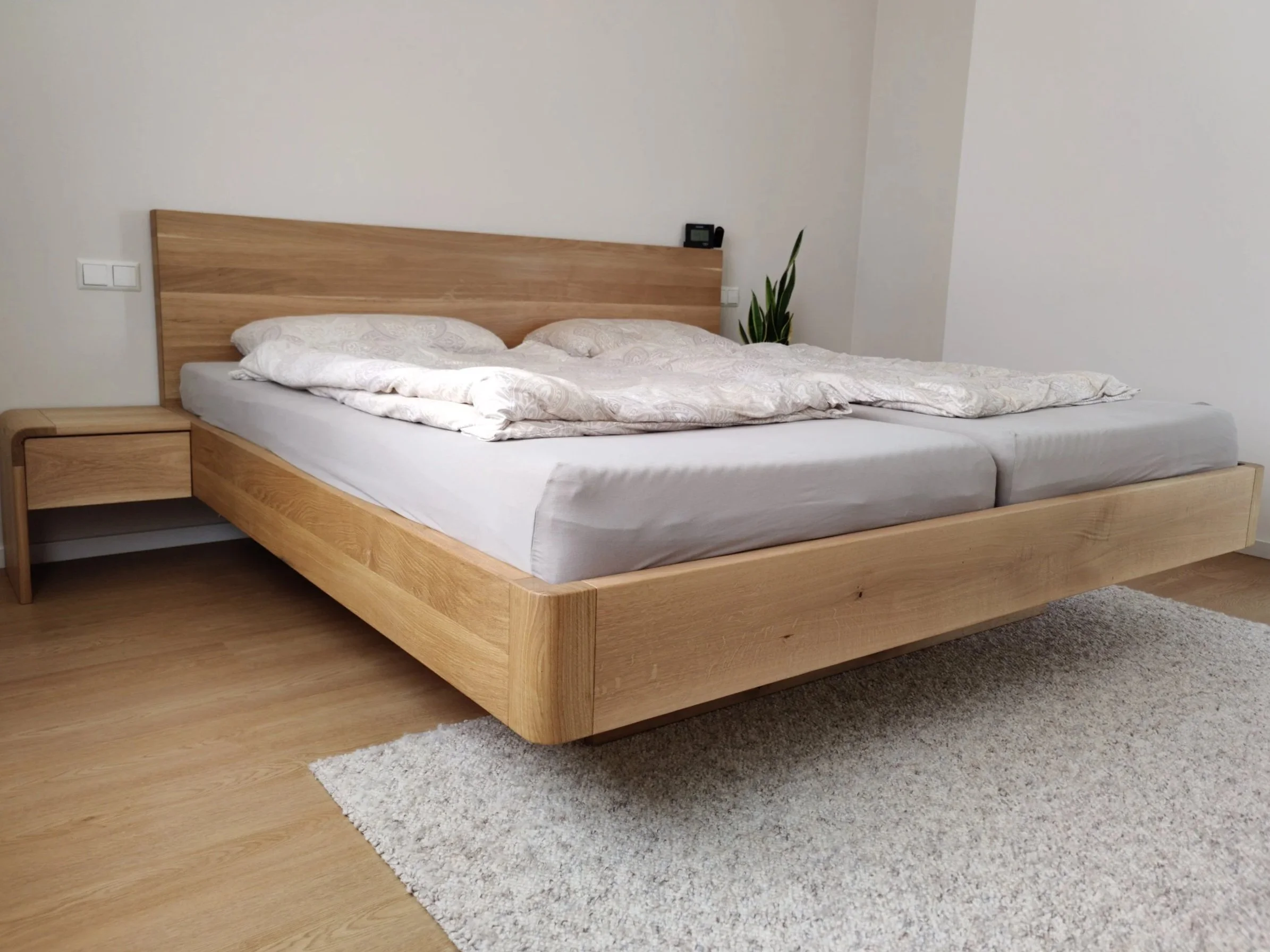 Ein modernes Schlafzimmer mit einem großen Holzbettrahmen und passenden Nachttischen, weißen Bettzeug und Kissen, einem Teppich und einer Topfpflanze in der Ecke.