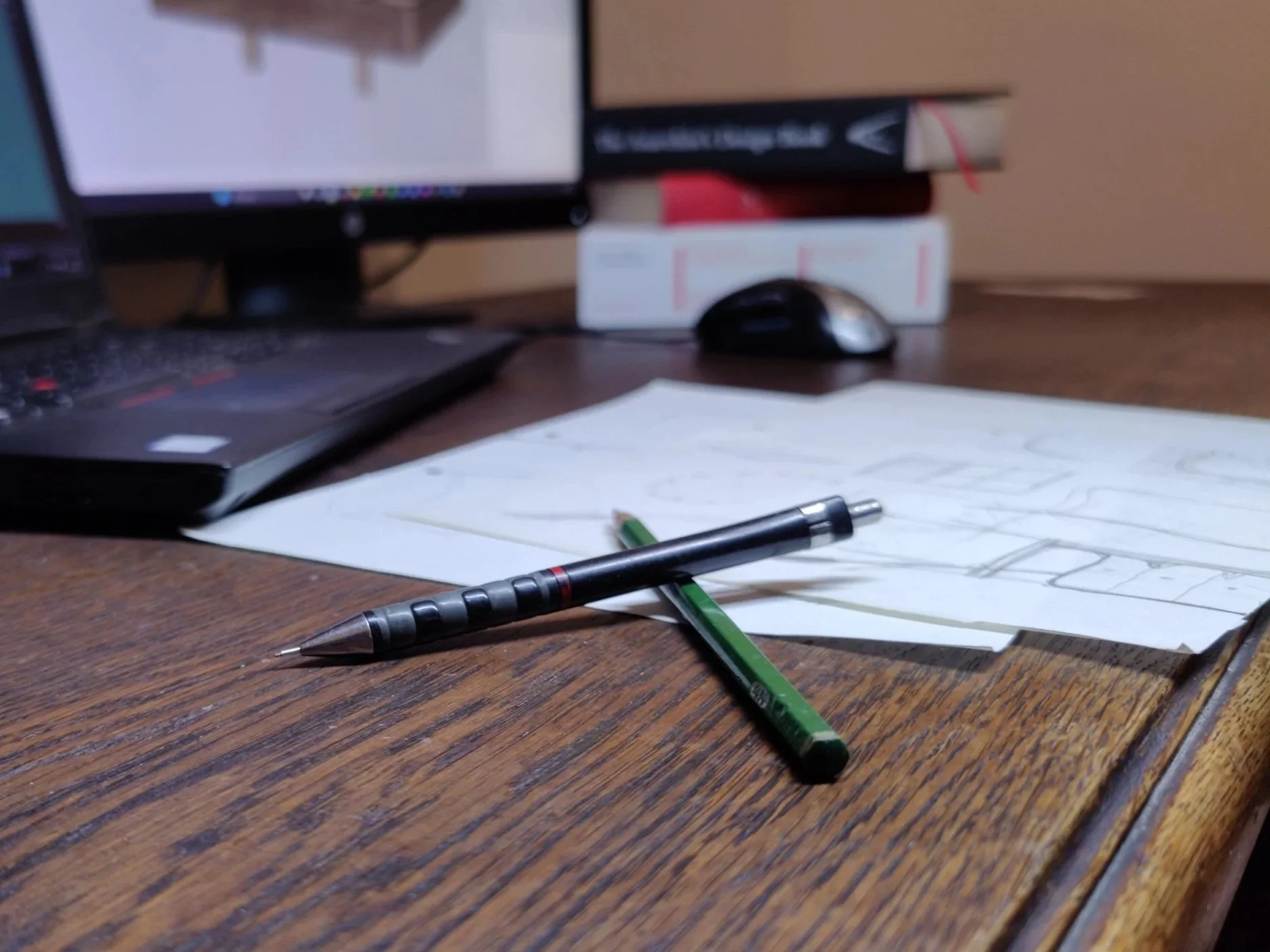 Schreibtisch mit Kugelschreiber, Zeichenstift, Papier mit Entwurf und einem Laptop, im Hintergrund Computerbildschirm, Bücher und Maus.
