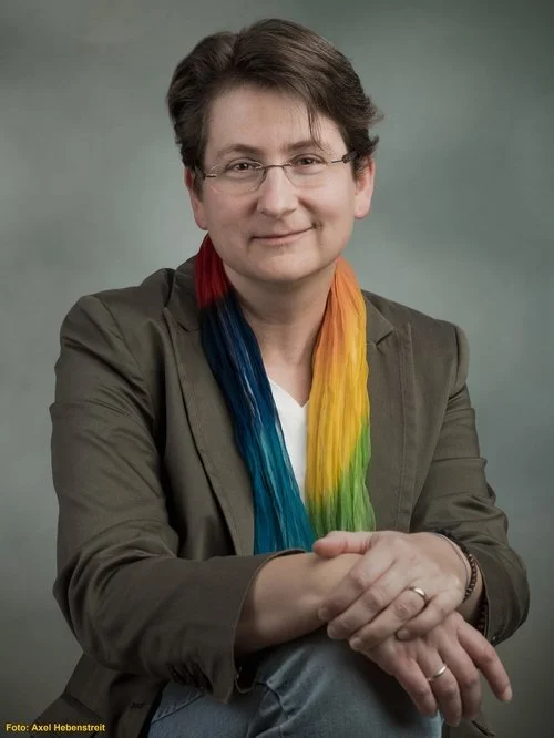 Eine Frau mit kurzen braunen Haaren, Brille und einem grauen Blazer, die einen bunten Regenbogen-Schal trägt, sitzt vor einem neutralen grauen Hintergrund.