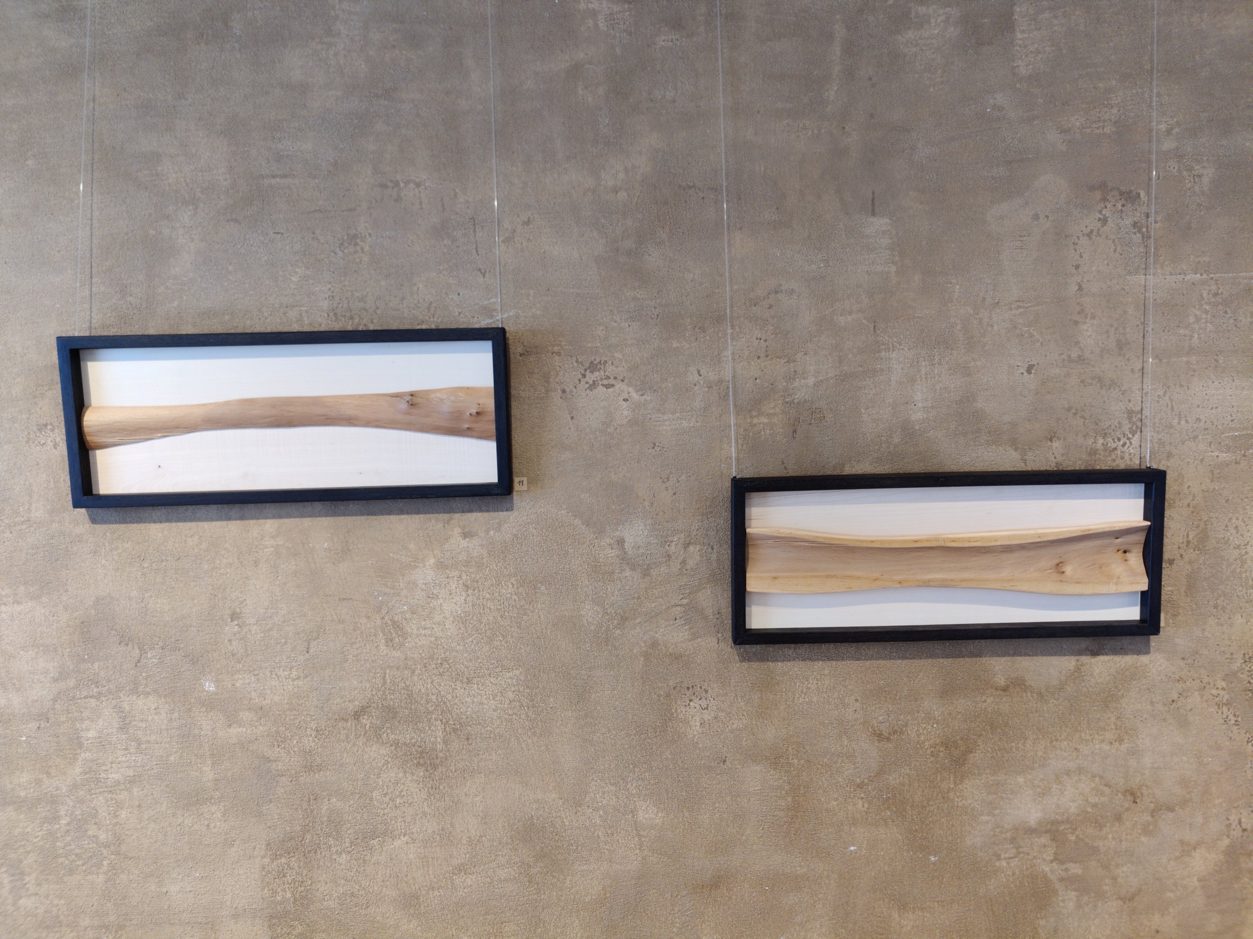 Zwei Kunstwerke mit Holzstücken in schwarzen Rahmen an einer beige Wand.