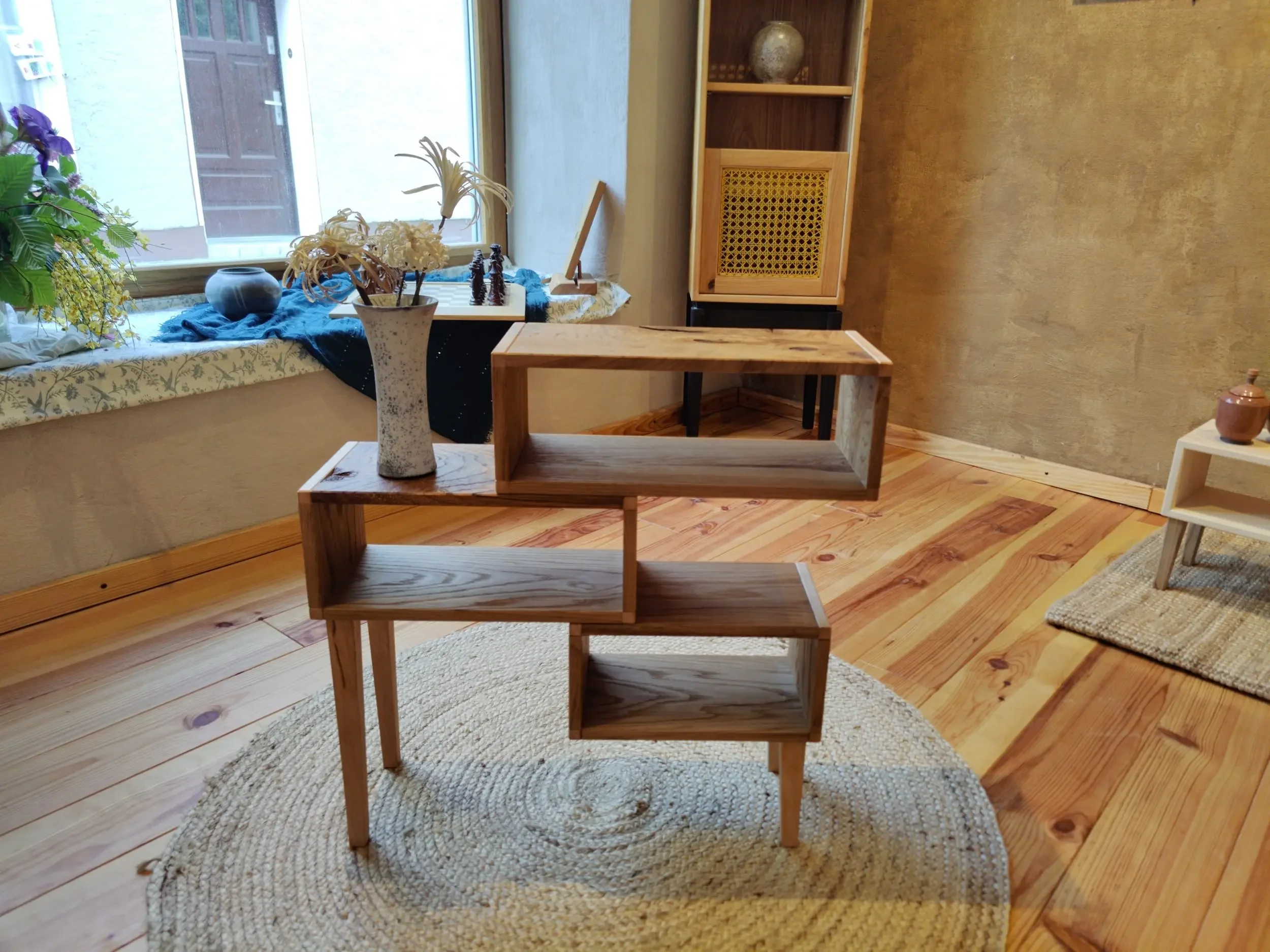 Ein Holzregal mit mehreren offenen Fächern in einem Wohnzimmer, auf einem runden geflochtenen Teppich, neben einem Fenster mit Blumenarrangement. Im Hintergrund eine Ecke mit Wandregal und Wohnzimmerdeko.