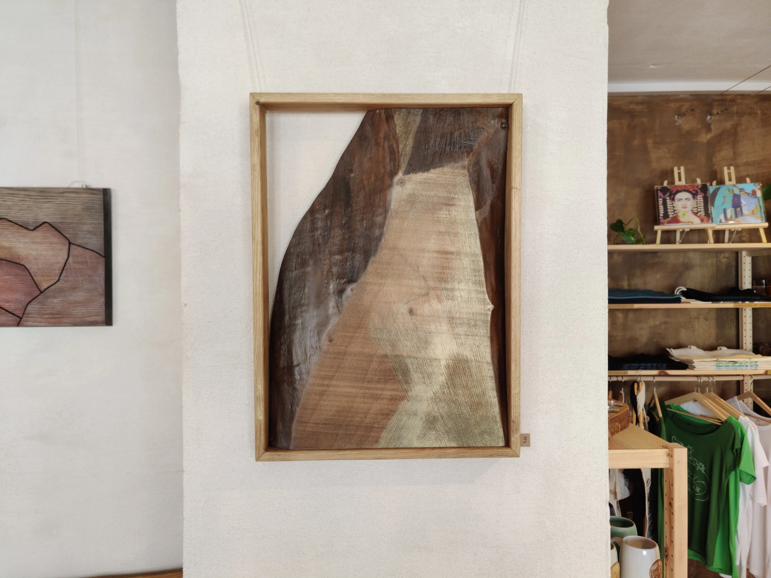 Holzkunstwerk mit holzfarbenen und braunen Holzstücken in einem schwarzen Rahmen, an einer weißen Wand in einem Raum mit verwinkeltem Bereich und Regalen mit Kleidung, Büchern und Kunstwerken.