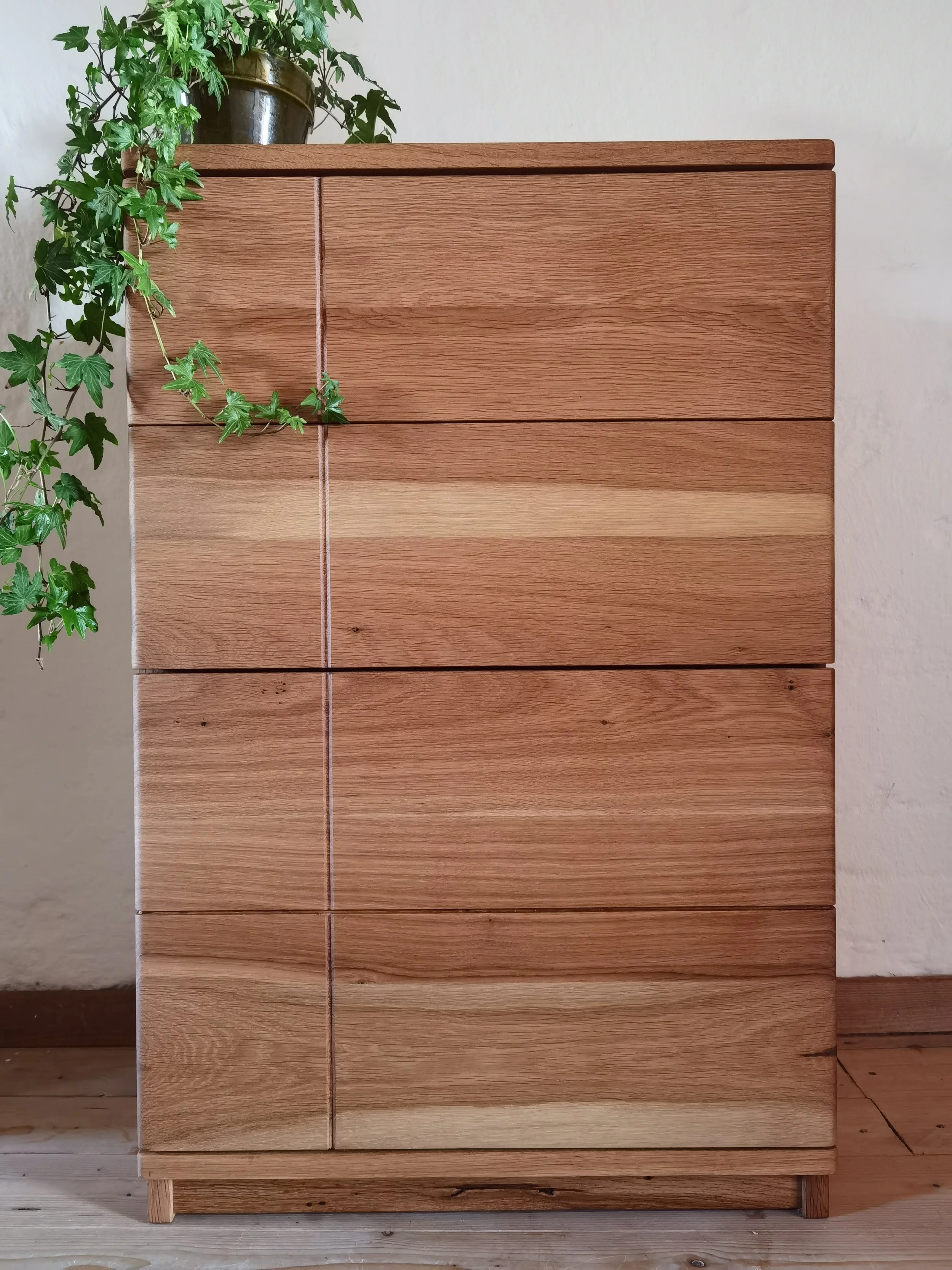 Ein Holzhocker mit drei Schubladen steht neben eine weiße Wand. Auf der Spitze steht eine grüne Zimmerpflanze in einem braunen Topf.