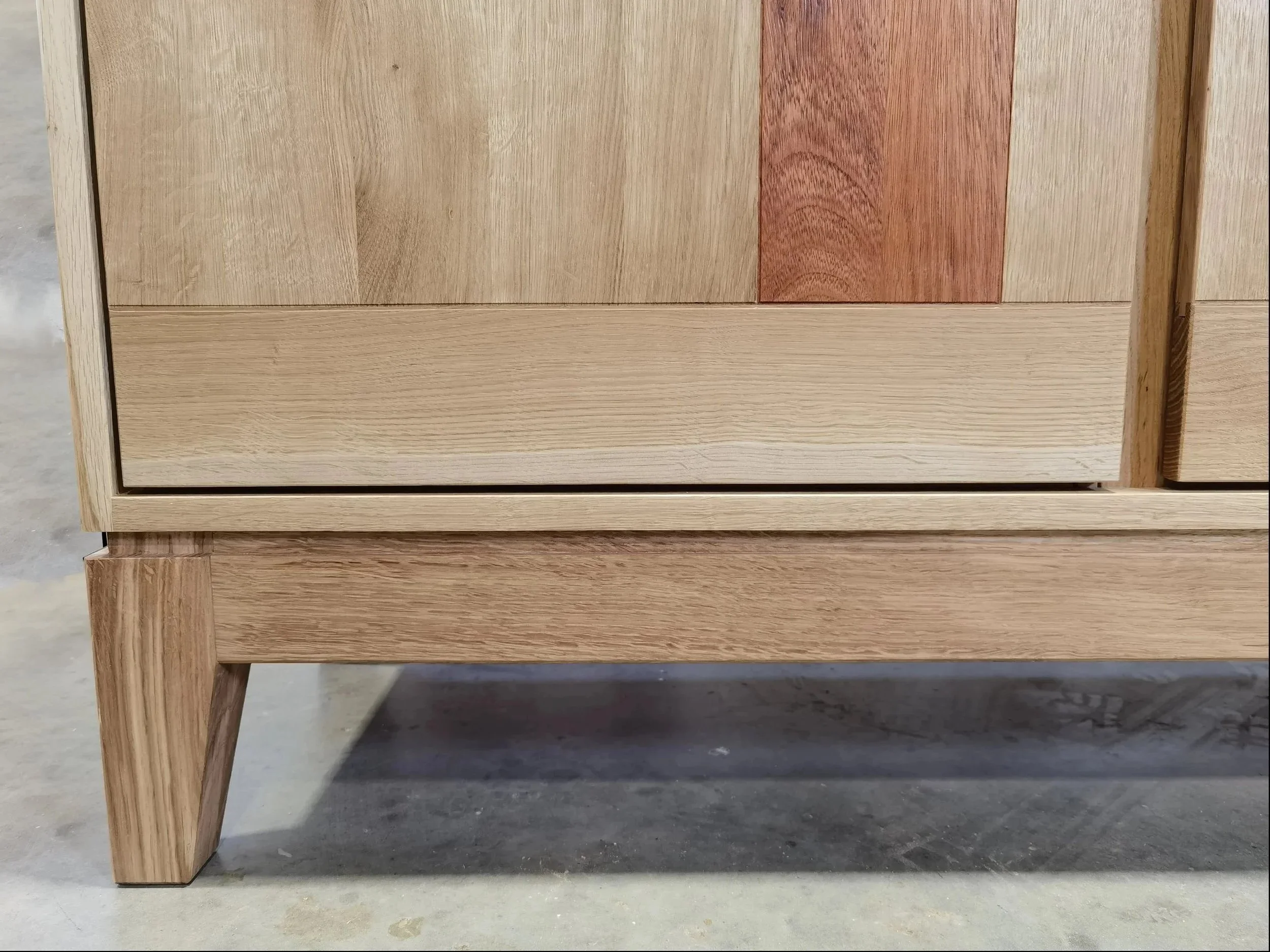 Nahaufnahme eines Holzmöbels, vermutlich eines Schreibtisches oder einer Kommode, aus verschiedenen Holzarten, auf einem Betonboden.