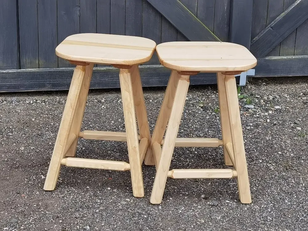 Zwei unbehandelte Holzhocker auf einem Kiesboden vor einem Holzzaun.