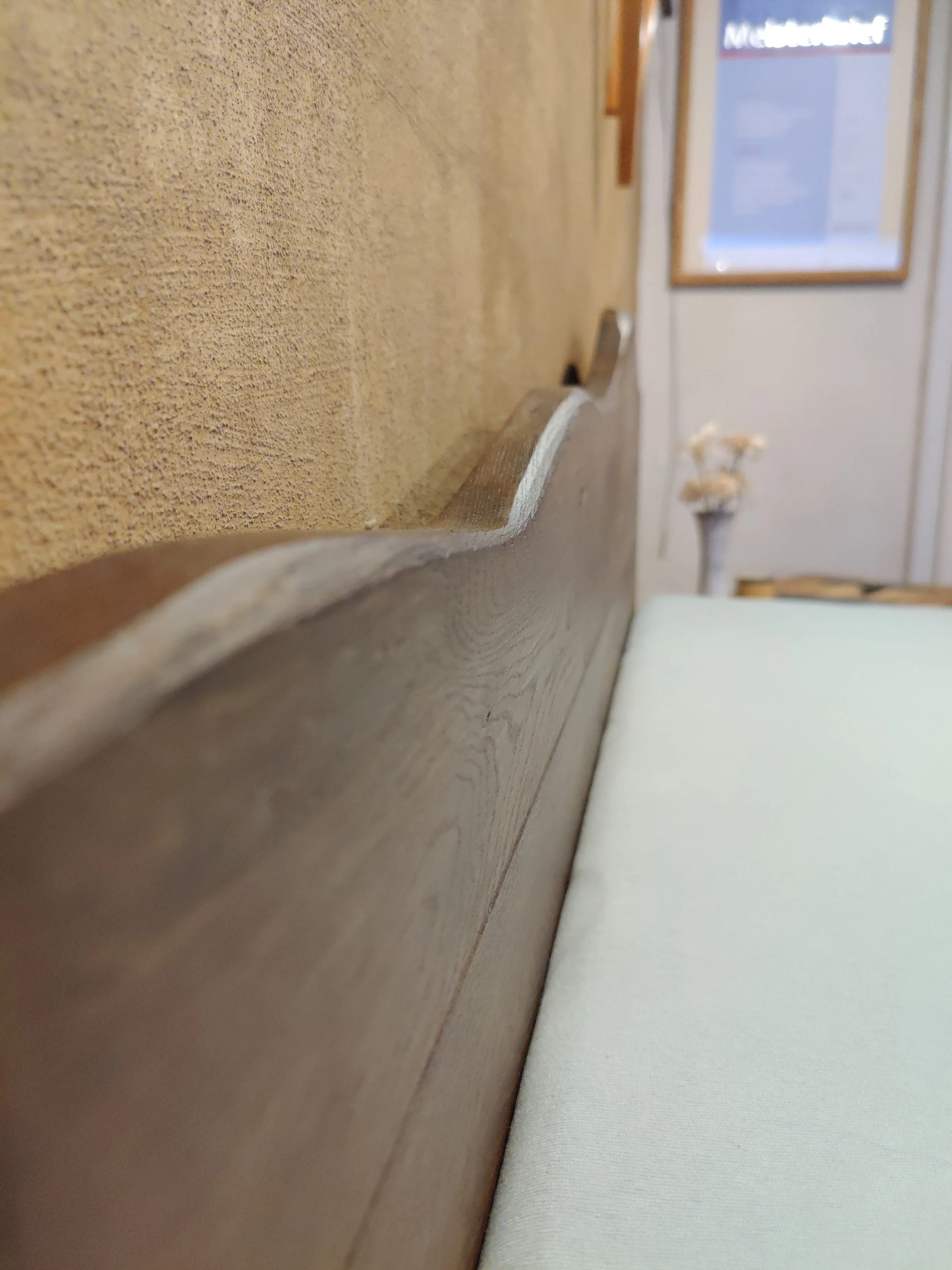 Nahaufnahme eines Holzkopfs mit ausgestrecktem Rücken und einem beige-weißen Kissen auf einer Bank, im Hintergrund eine Wand mit einem Rahmen und einer Vase mit weißen Blumen.