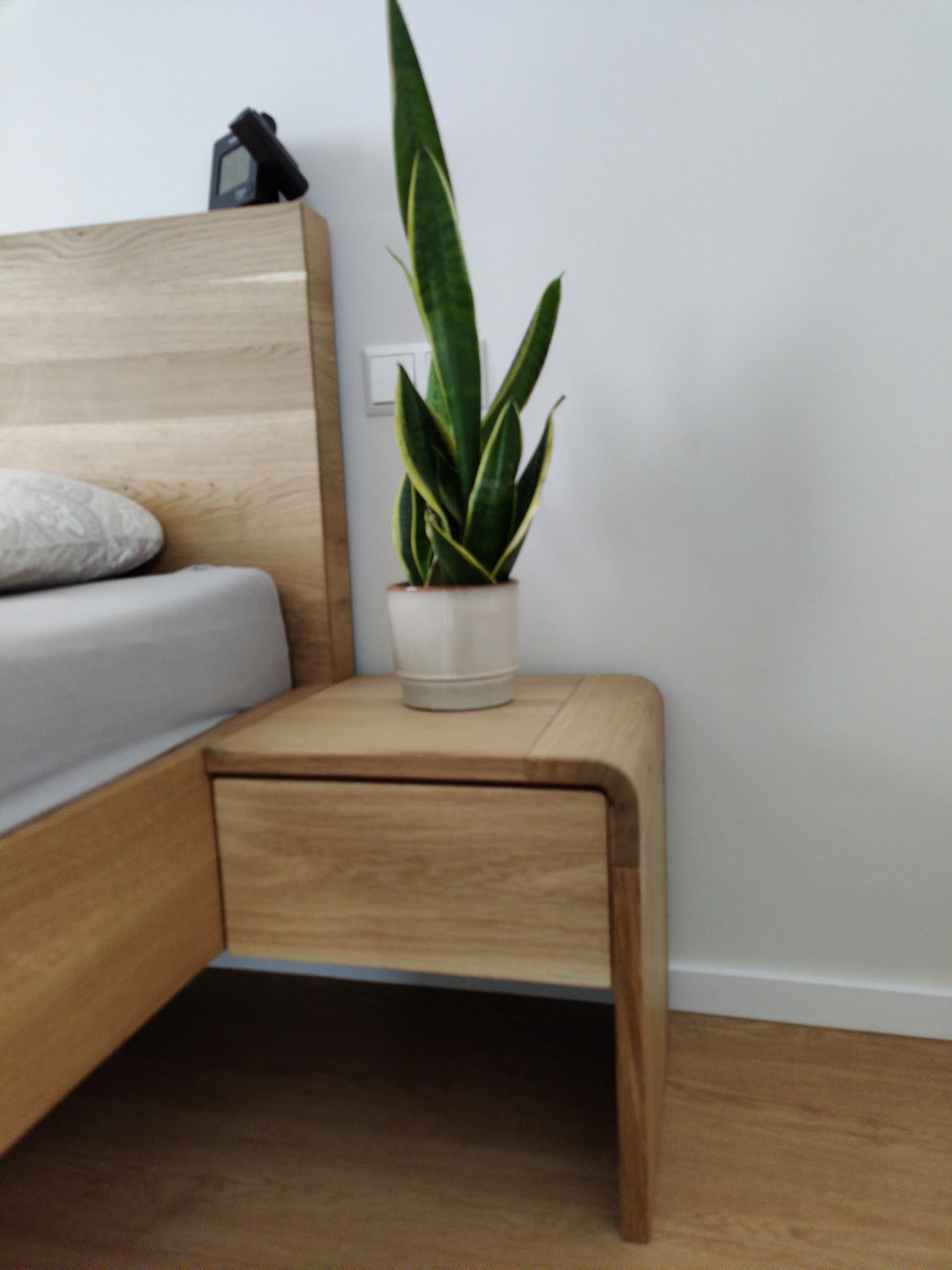 Ein Holznachttisch mit einer Pflanze auf einem weißen Topf, neben einem Bett mit weißer Bettwäsche und einem Kopfteil aus Holz.