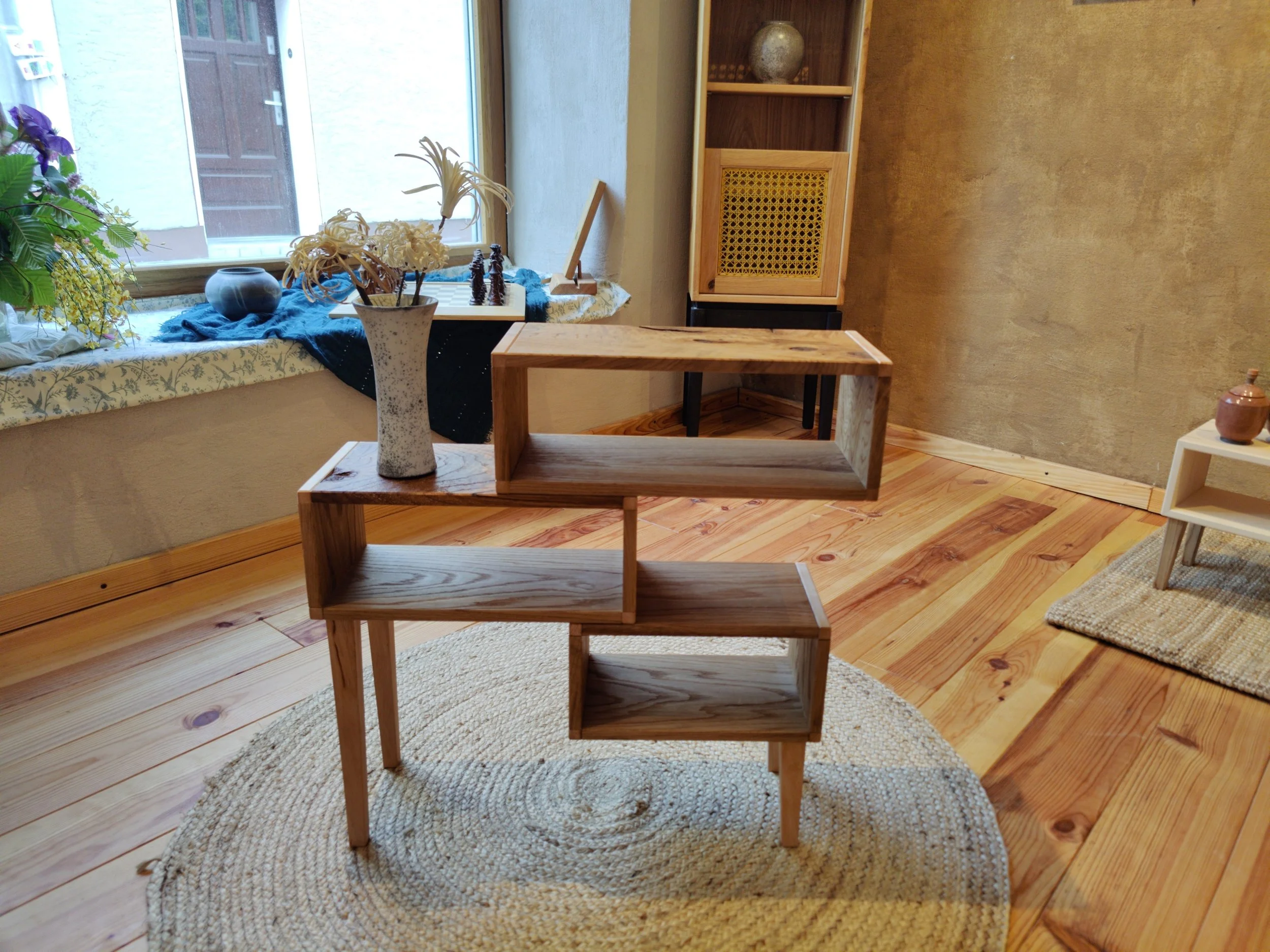 Ein stilvoller Raum mit Holzfußboden, einer Naturholzkiste in der Mitte, dekorative Gegenstände auf der Fensterbank und einem weißen Regal im Hintergrund.