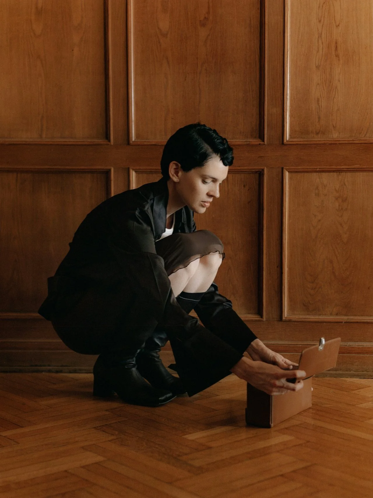 A woman with short dark hair, dressed in black, crouching in front of wood-paneled wall, placing an envelope into a wooden mailbox on the floor.