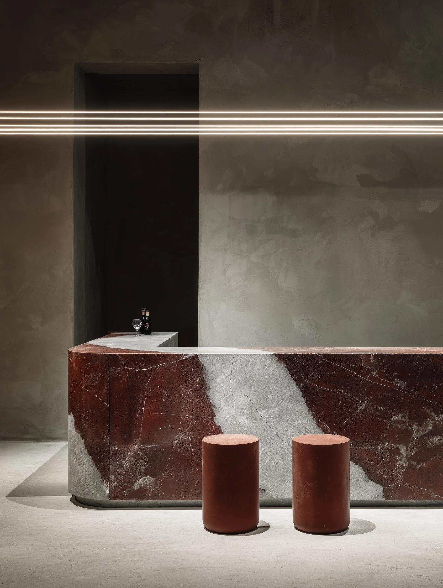 Modern bar with a curved red marble counter, two red cylindrical stools, and a small table with bottles and glasses in a minimalistic, dimly lit setting.