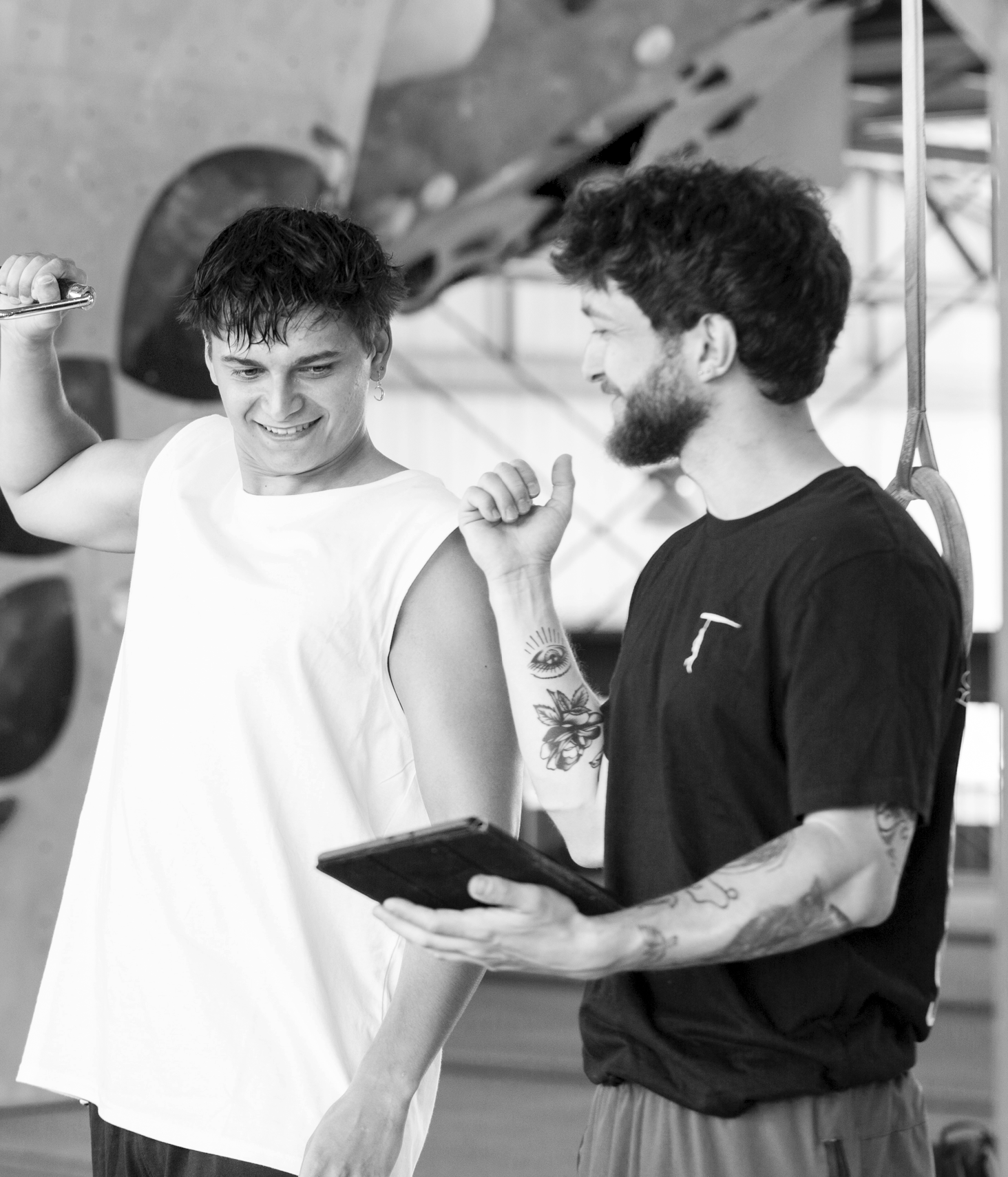 Zwei Personen, eine junge Frau und ein junger Mann mit Tattoos, lachen und sprechen sich in einem Klettergym an