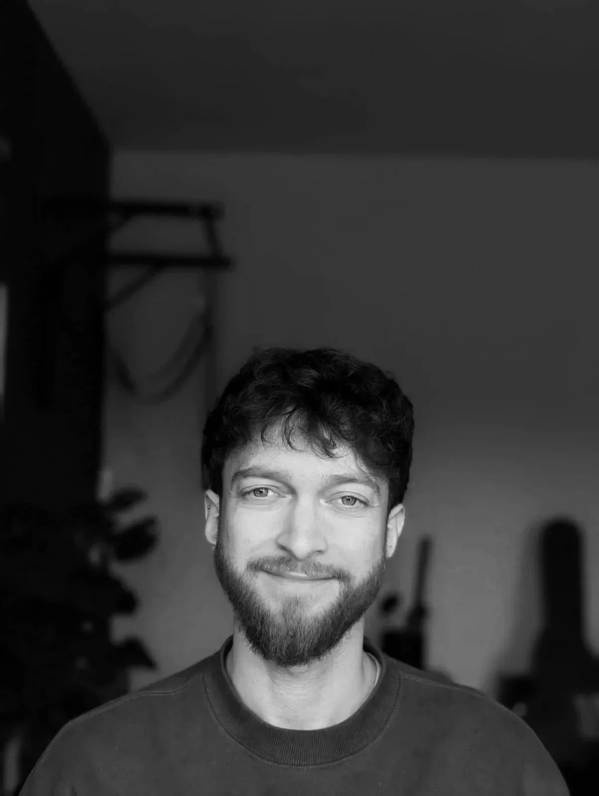 Schwarz-Weiß-Foto eines lachenden jungen Mannes mit dunklem, lockigem Haar und Bart, im Hintergrund unscharfe Möbel.