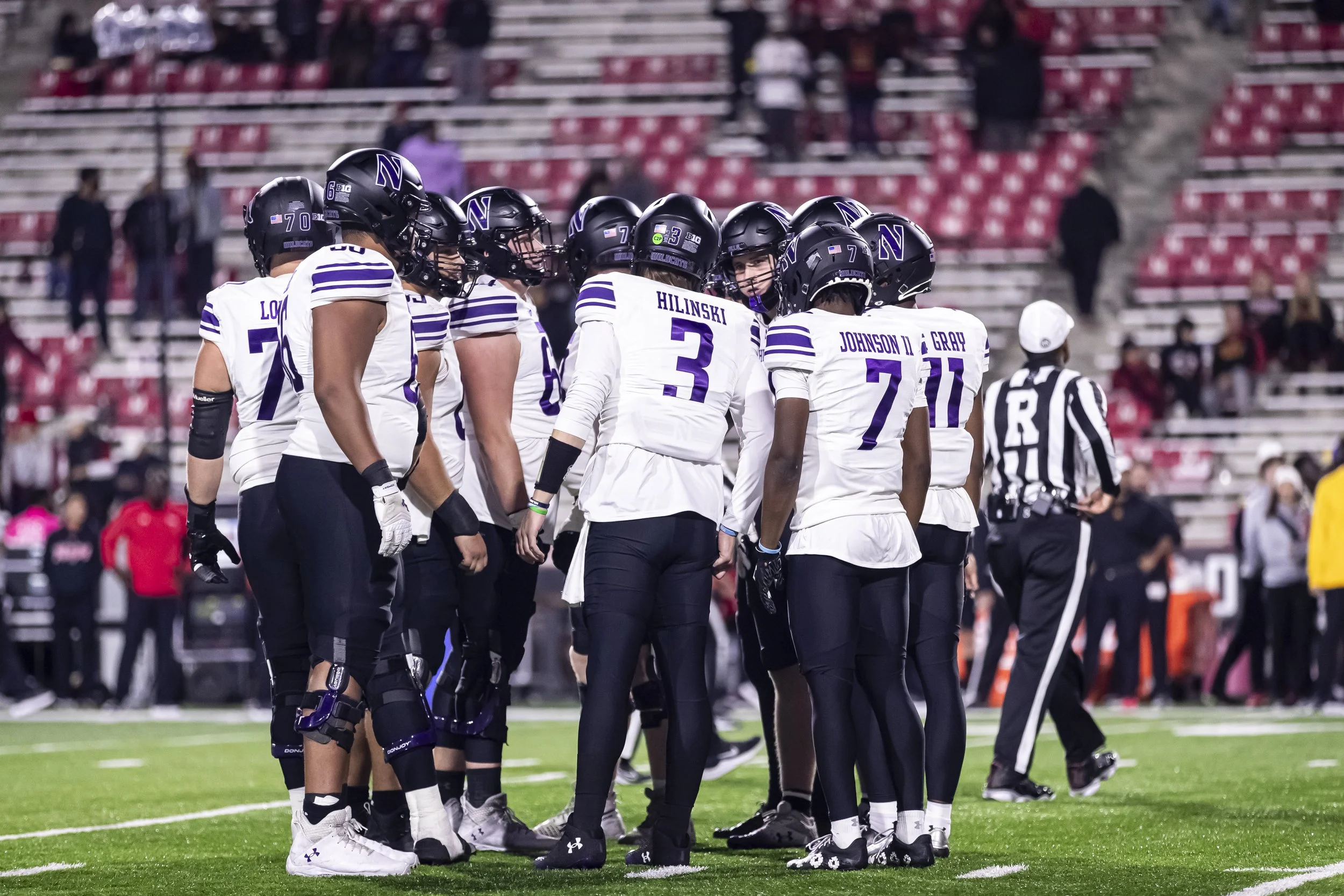 A group of American football players in white and purple uniforms huddle on the field during a game, with a referee nearby and spectators in the stands.