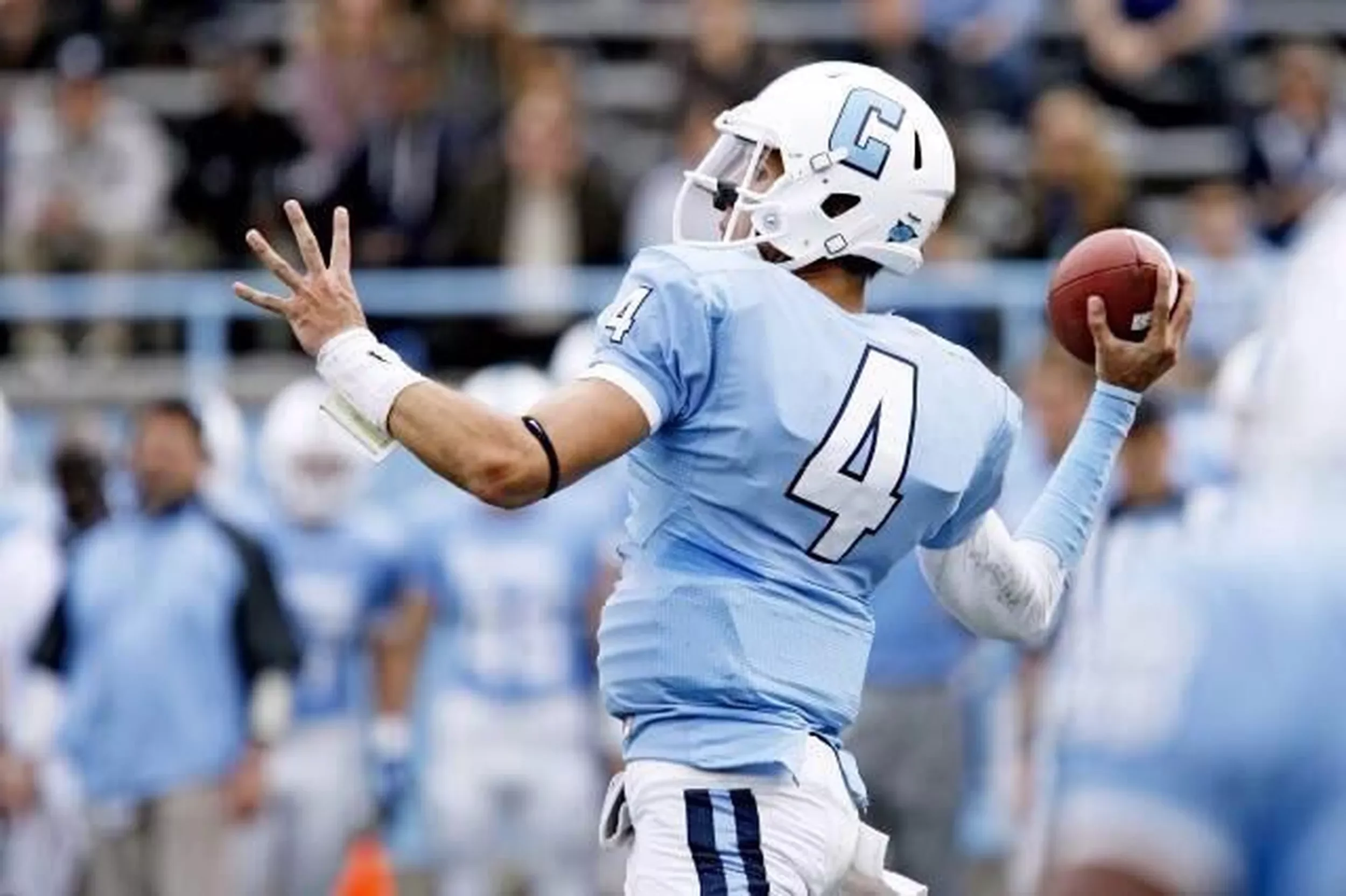 A football player wearing a light blue jersey with the number 4 and a white helmet is in motion, holding a football in his right hand, with an audience in the background.