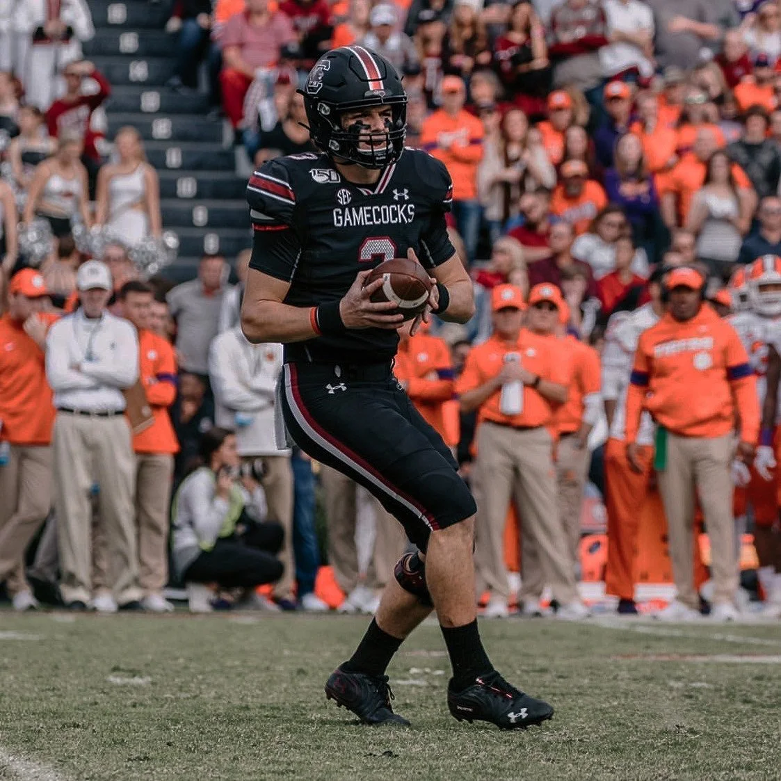 A football player in a black uniform with red and white accents, holding a football and preparing to run. The uniform has the word 'GAMECOCKS' on it, and the player is wearing a helmet and cleats. Spectators and staff in orange and khaki clothing are visible in the background.