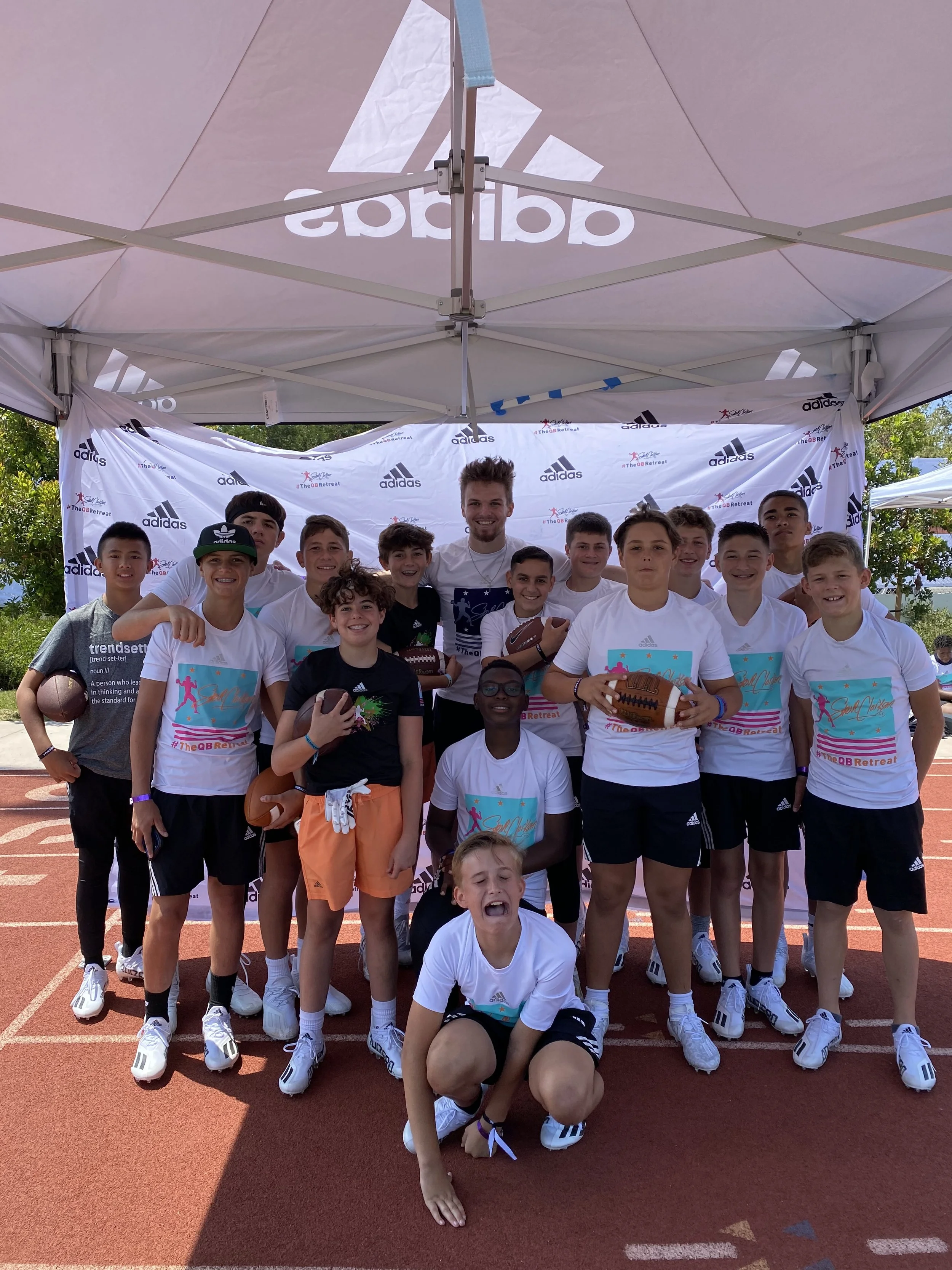 Group of young athletes and coach posing under a white Adidas tent on a running track, holding footballs, shirts, and wearing sports gear.