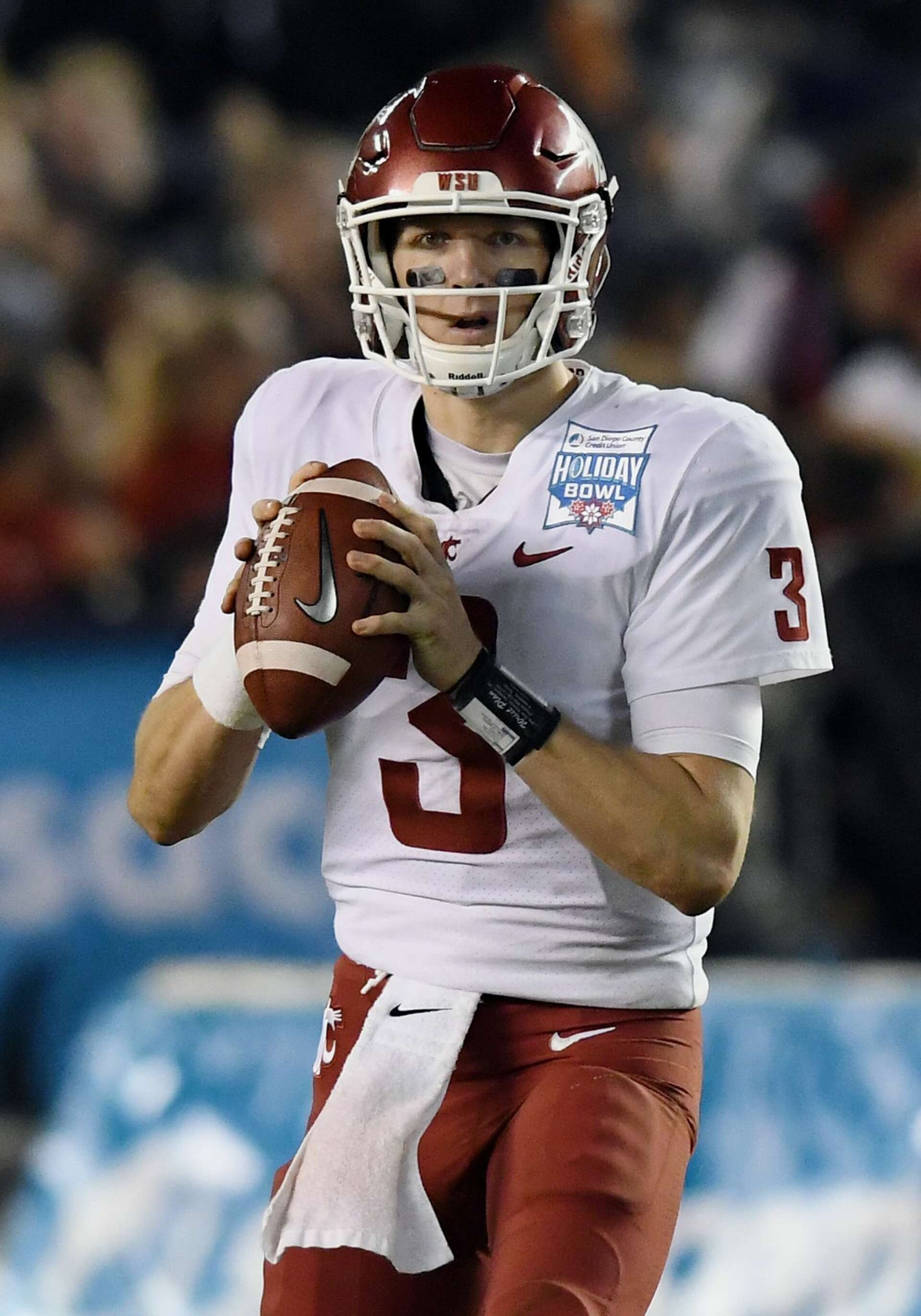 A football player in a white and red uniform holding a football, wearing a helmet, during a game. The uniform has a patch that says 'Holiday Bowl'.