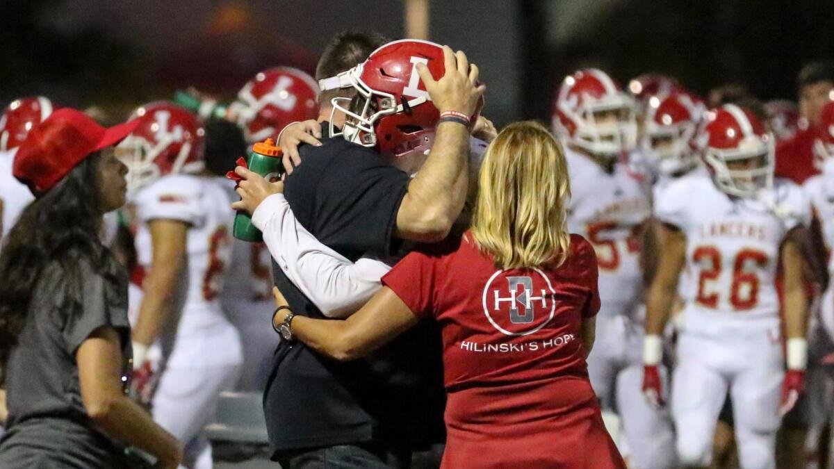 A football coach or staff member embracing a player during a game, with other players in white and red uniforms in the background, and a woman in a red shirt watching nearby.