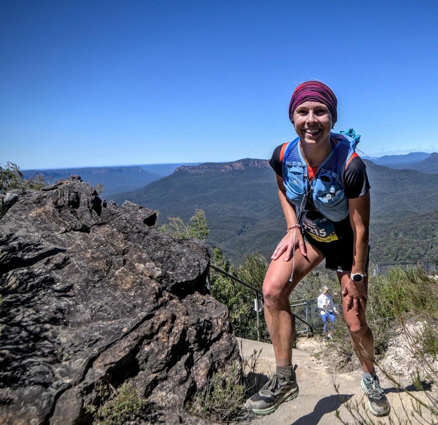 A woman smiling and in running gear, including a headband and hydration vest, climbing on a rocky trail with scenic mountains and clear blue sky in the background.