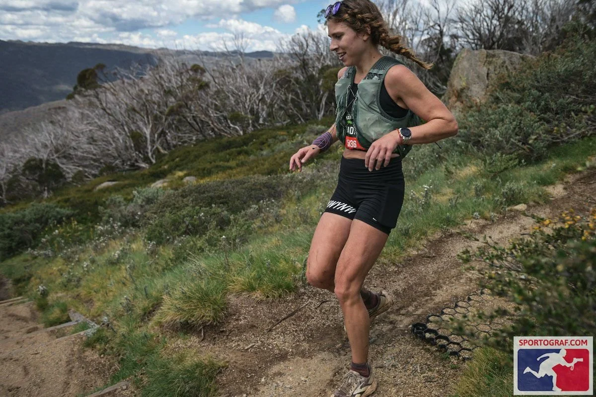 A woman trail running on a mountain path with vegetation and distant mountains in the background.