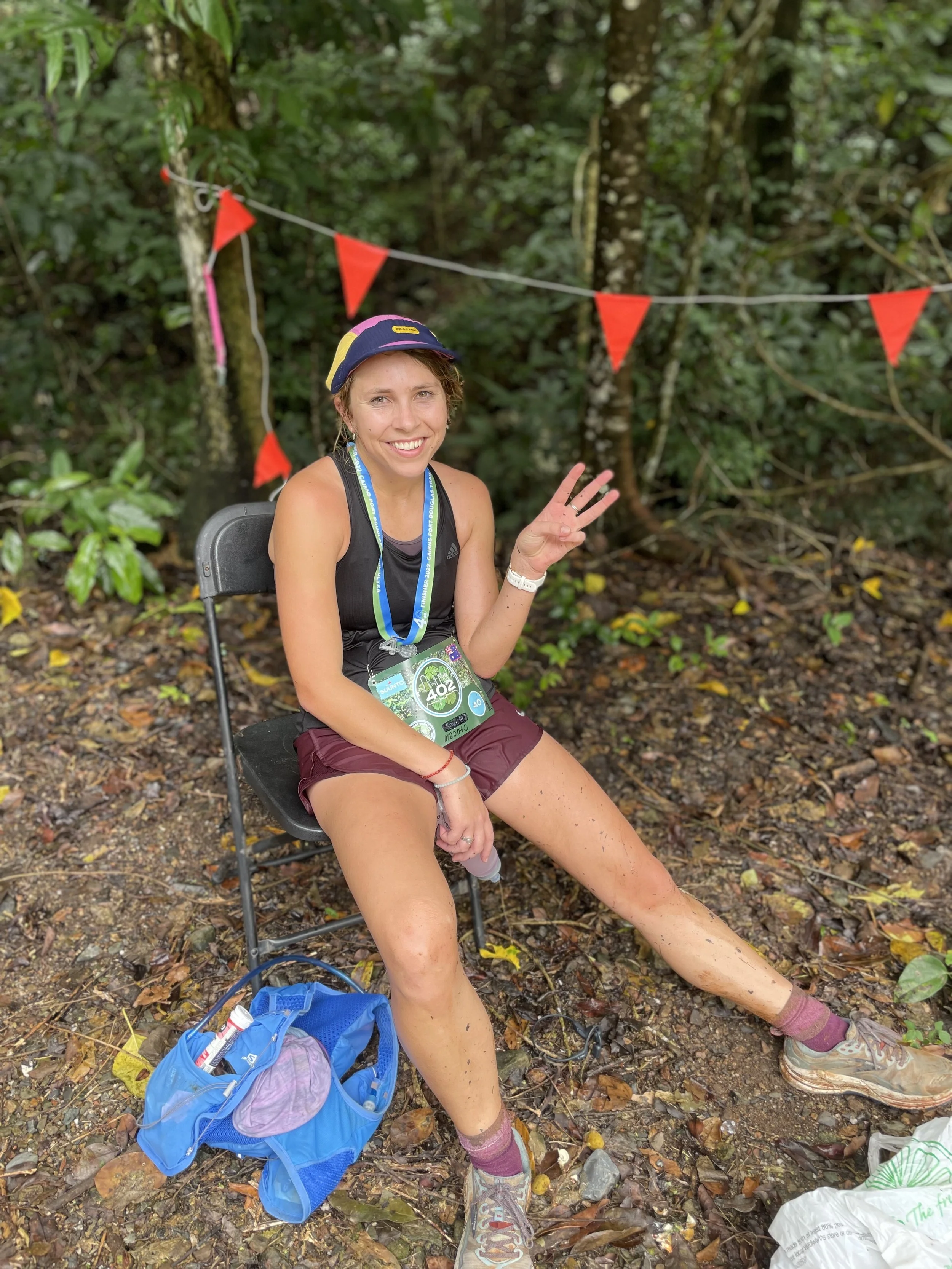 A woman sitting on a folding chair in a forest, wearing running gear and a cap, smiling and waving, with a race bib on her chest and a blue backpack on the ground nearby.