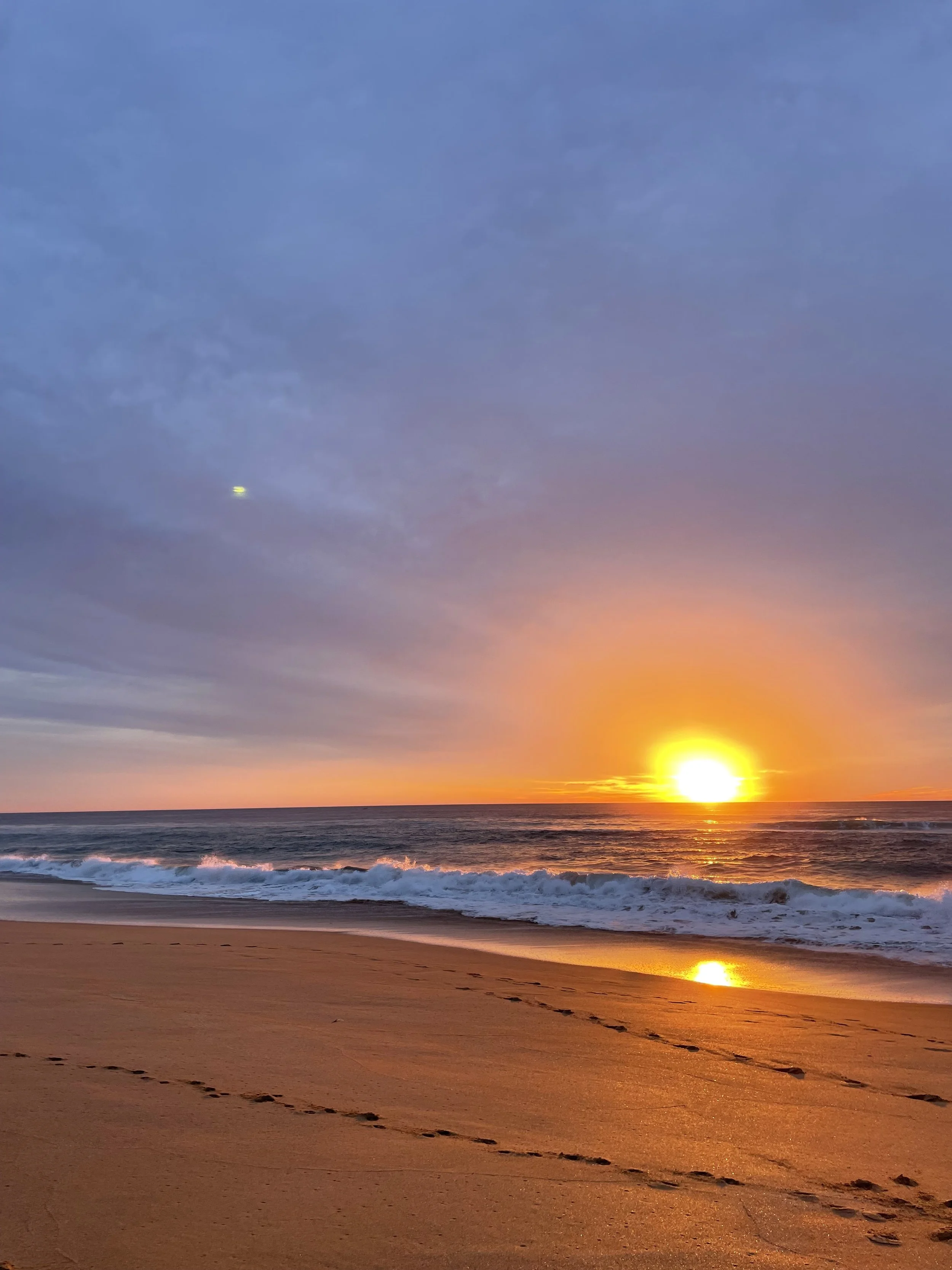 Sunset over the ocean with waves crashing onto the sandy beach, footprints in the sand, and a cloudy sky with a small bright object.