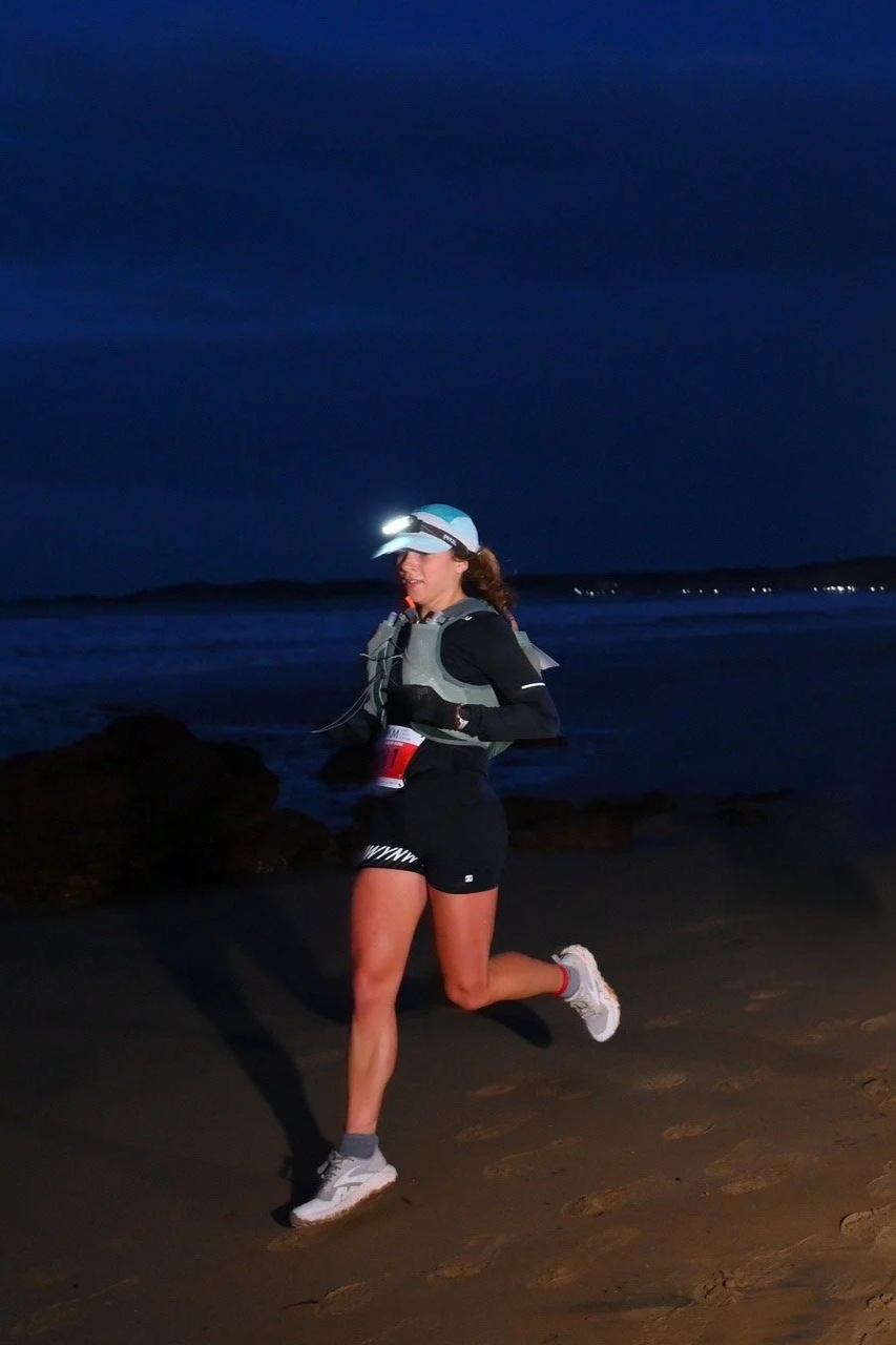 A woman running on the beach at night with a headlamp, wearing athletic gear and a hydration vest.
