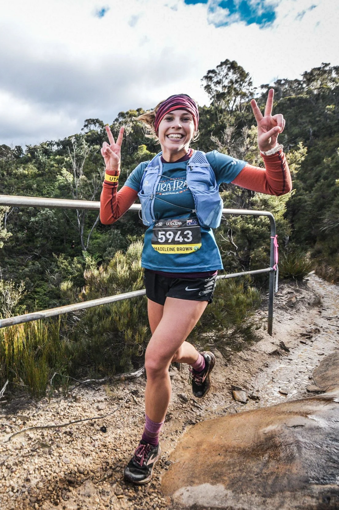 Female trail runner making peace signs while running on a dusty dirt trail with a metal railing, forested area, and cloudy sky in the background.