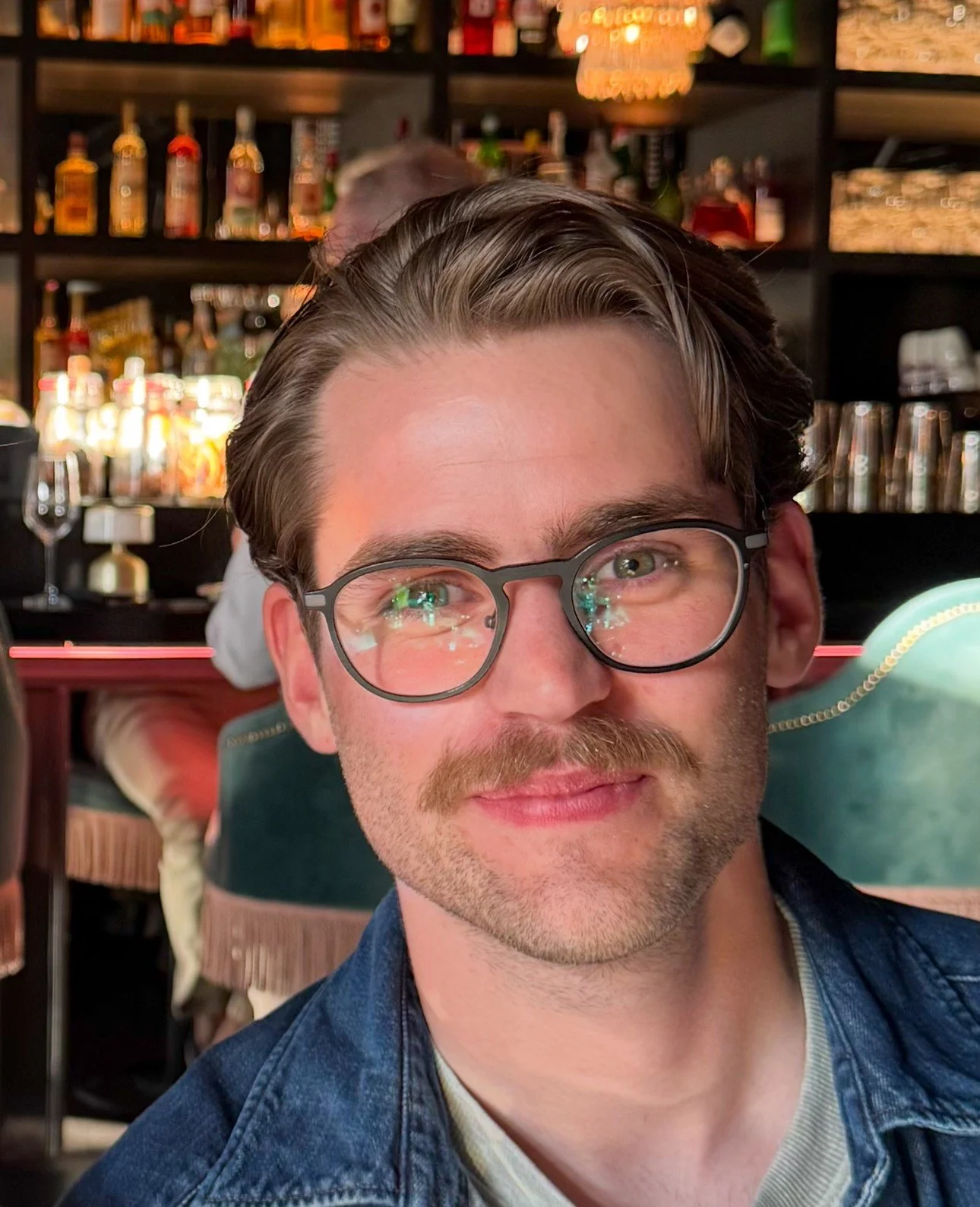 A smiling man with glasses and a mustache in a bar or restaurant setting with shelves of alcohol bottles in the background.