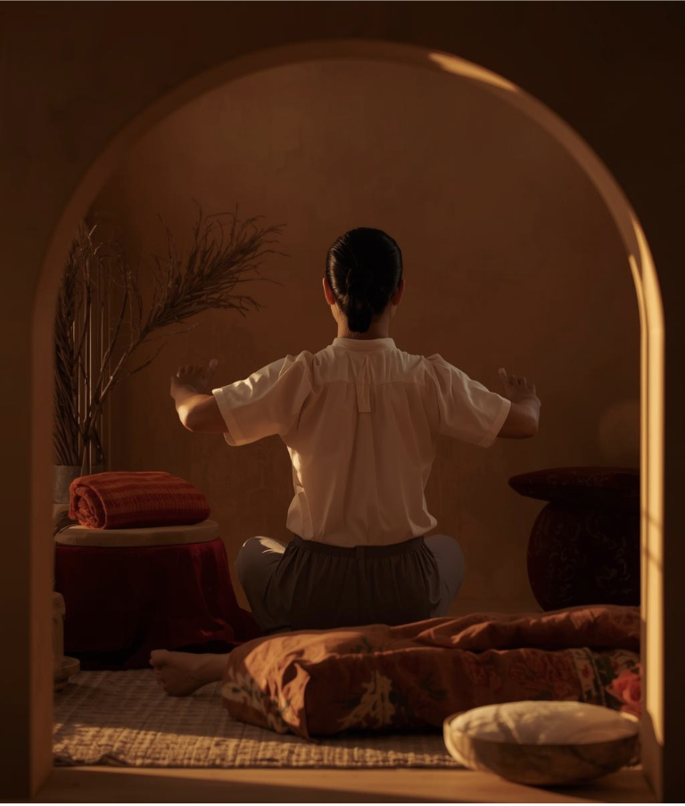 Person sitting cross-legged in a room, facing away, practicing yoga or meditation during sunset, with warm lighting, near cushions and a decorative vase.