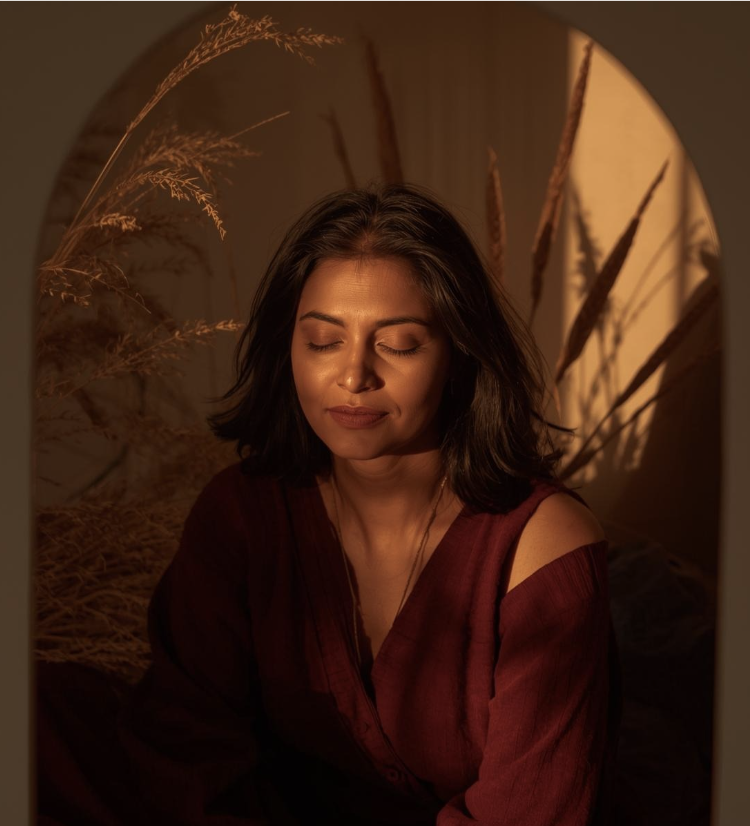 A woman with dark hair and closed eyes, sitting indoors with warm lighting, wearing a burgundy top with cut-out shoulders, surrounded by dried plants.