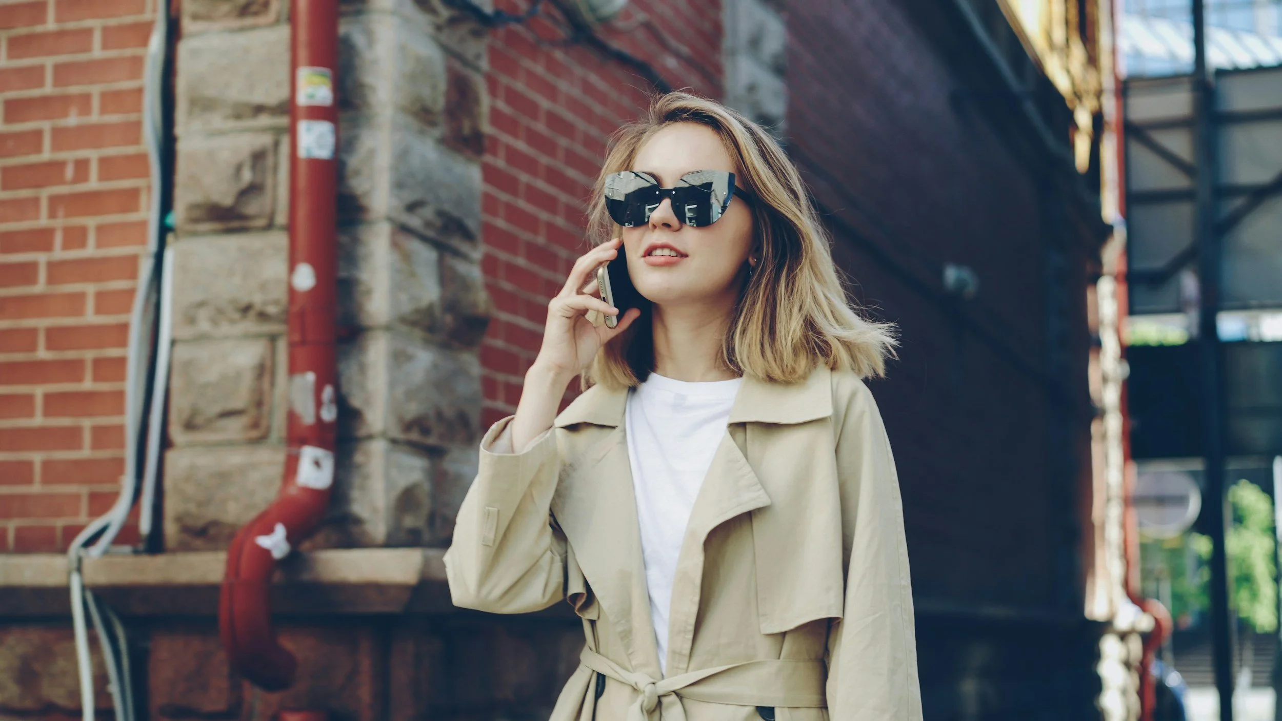 A woman with shoulder-length blonde hair wearing large black sunglasses, a beige trench coat, and a white T-shirt, talking on a cellphone on an urban street with brick buildings.