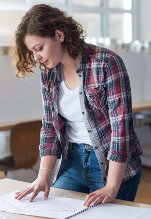 Young woman with curly hair in a plaid shirt looking at papers on a table in a bright room.