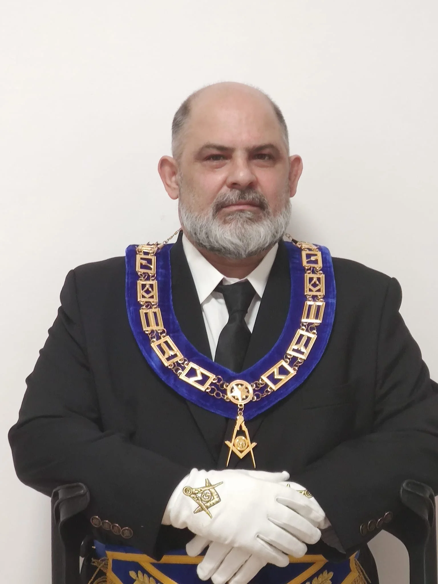 Uomo con barba, vestito elegante con giacca nera e cravatta nera, indossa un collare rituale ricco di dettagli dorati e blu, con un simbolo a forma di stella e strumenti di disegno, e guanti bianchi con dettagli dorati, seduto contro uno sfondo bianco.