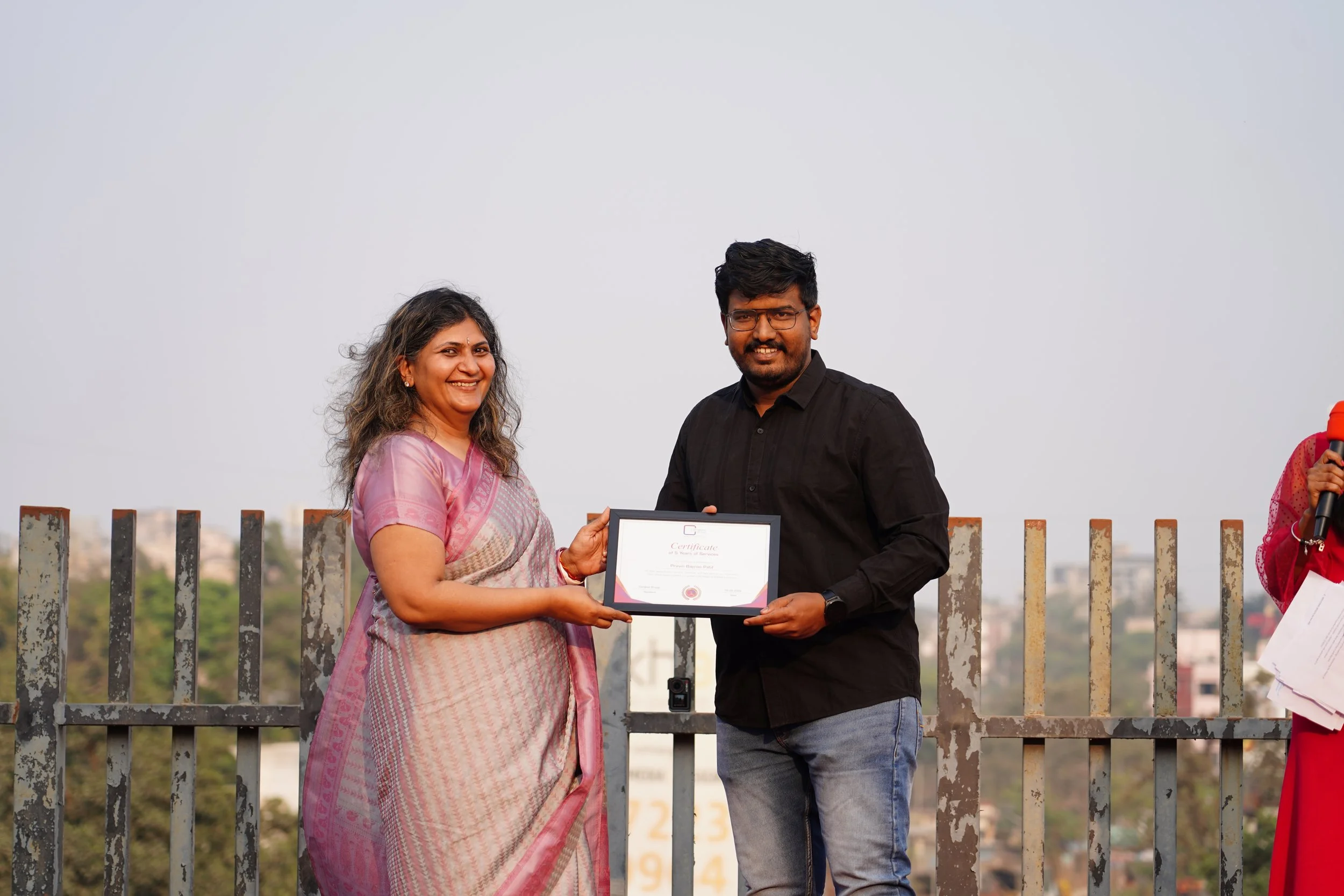 A woman in traditional attire is presenting a certificate to a man in a black shirt and jeans on a rooftop with a cityscape in the background during a daytime outdoor ceremony.