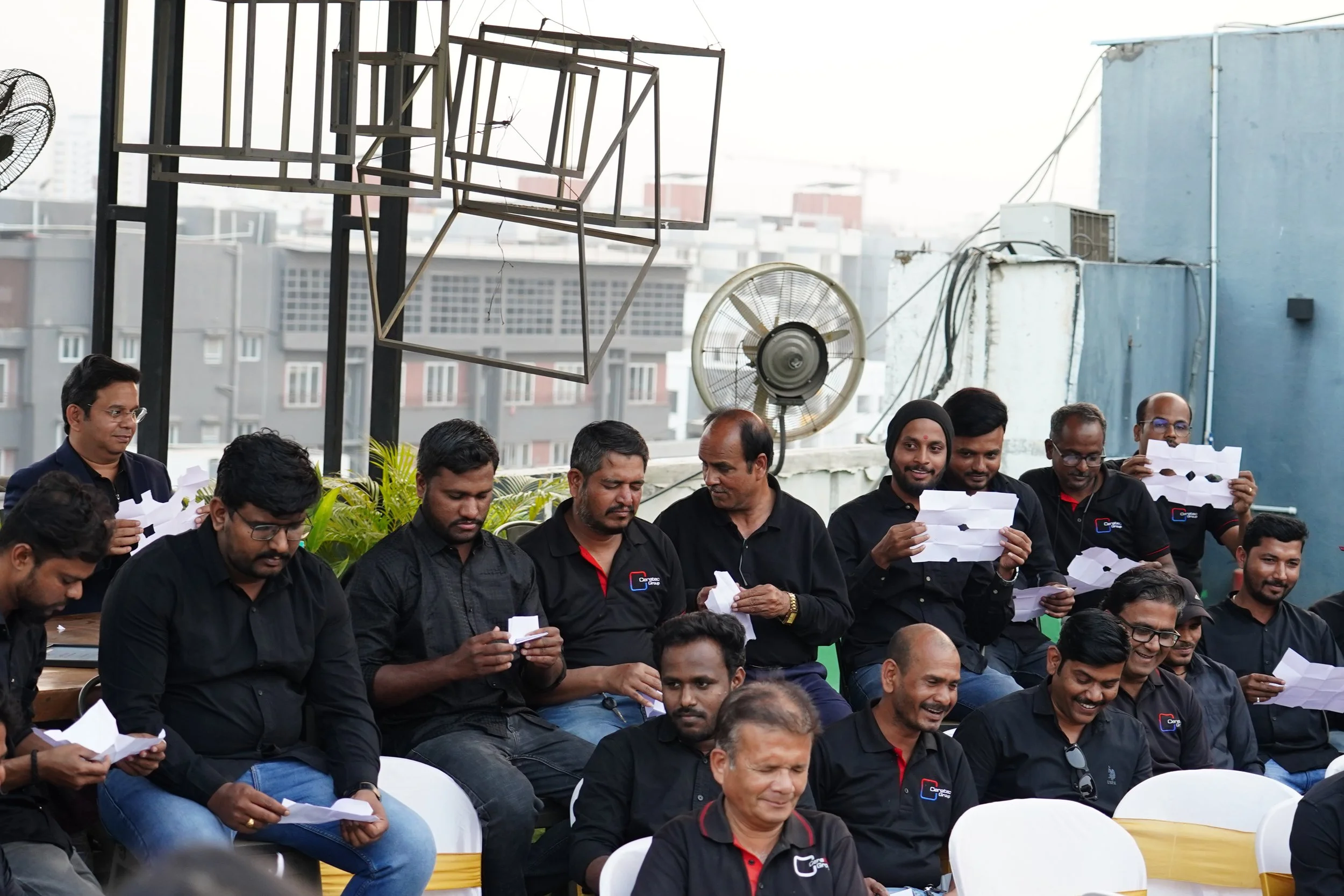 Group of men seated and standing on a rooftop, most wearing black shirts, some holding paper airplane crafts and smiling.